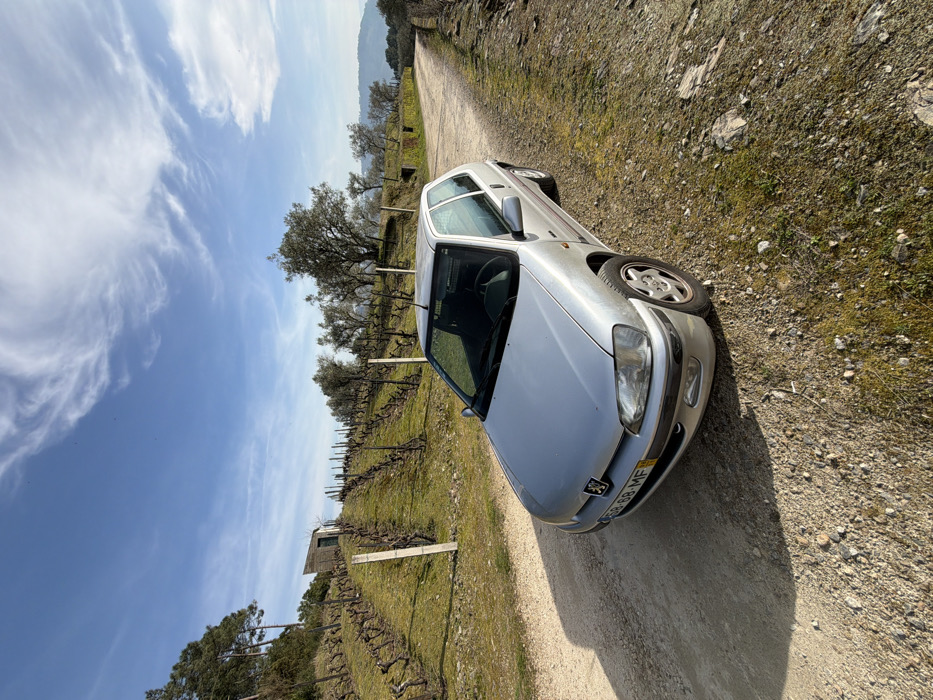 A silver Peugeot 106 is parked on a dirt road next to a vineyard. The car faces the camera, with the license plate partially visible. The vineyard has rows of grapevines and some trees in the background. The sky is blue with scattered clouds, and the overall setting appears to be rural.