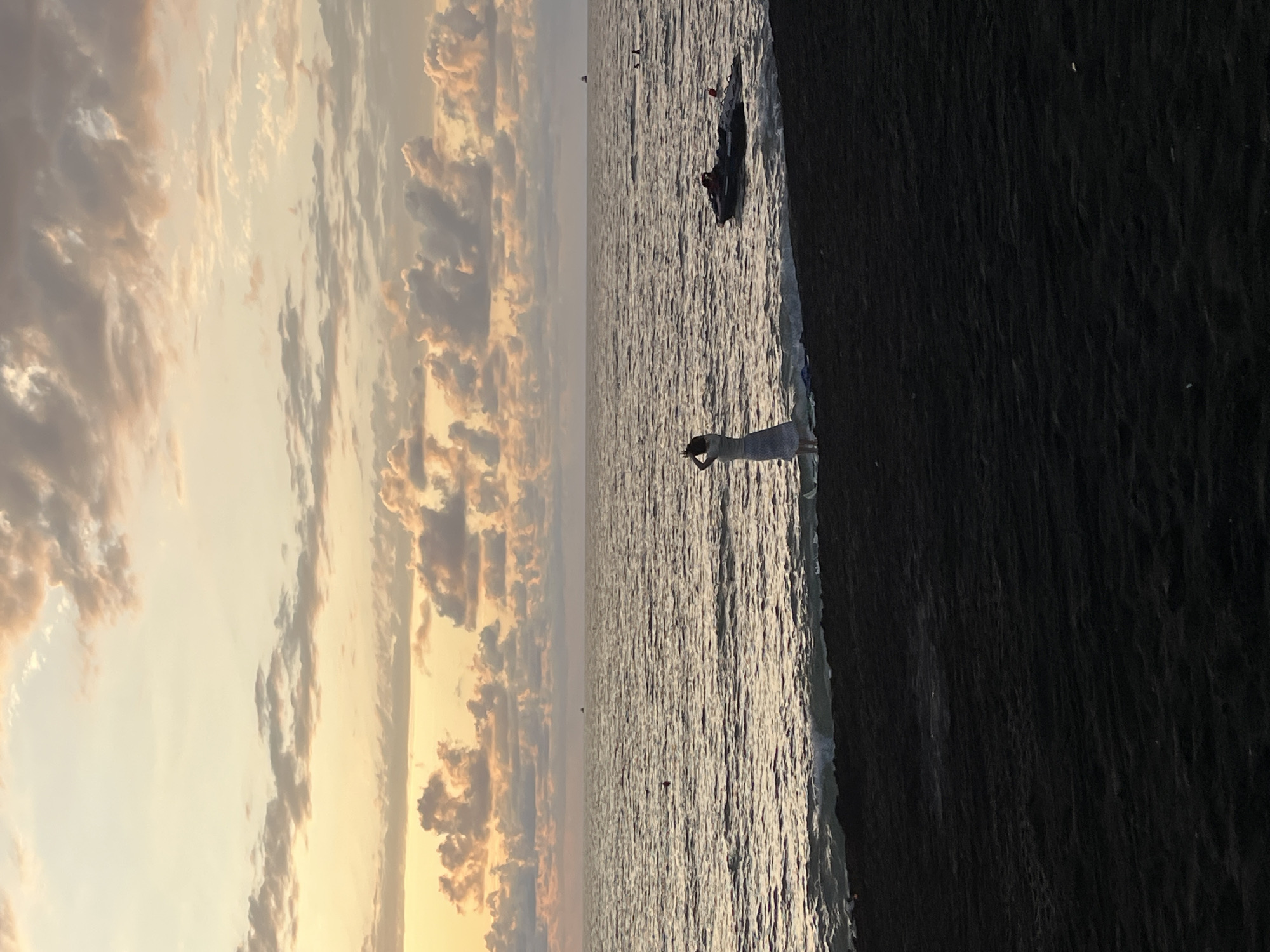 A solitary woman stands on the dark sand of Ureki Beach, Georgia, facing the sea as gentle waves lap at the shore. She is dressed in a light-colored, ankle-length dress and her hands are raised to her head. In the distance, a jet ski is visible on the water. The sky above is filled with soft, pastel-colored clouds, indicative of either sunrise or sunset. The scene conveys a sense of peaceful solitude and natural beauty.