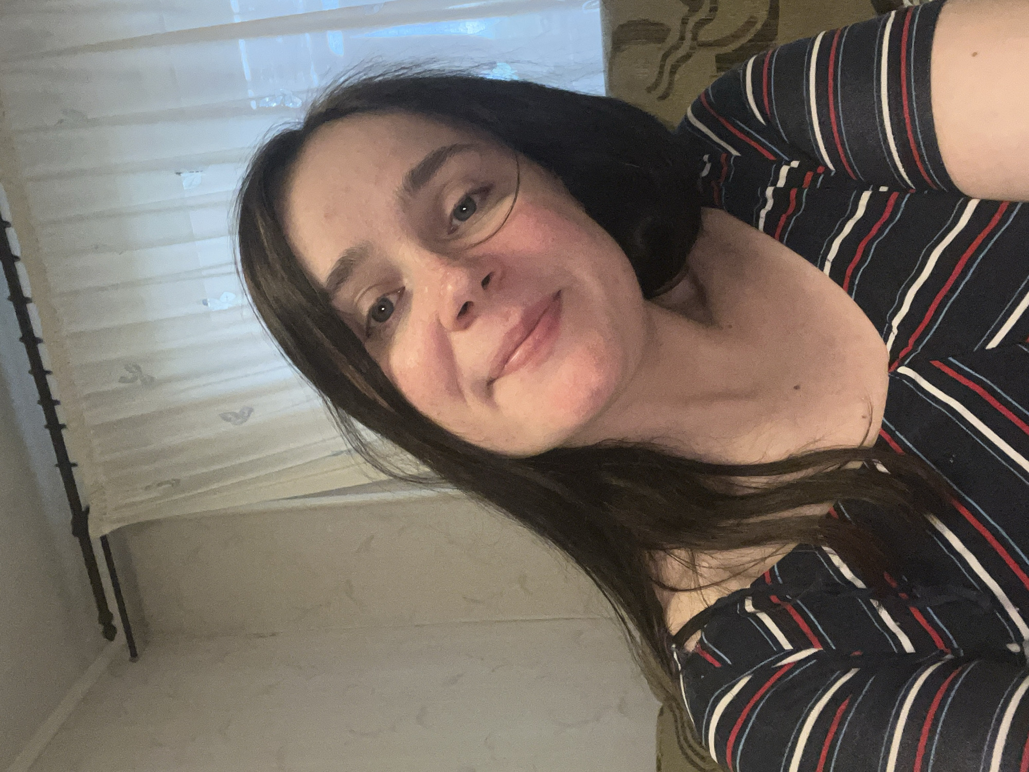 This is a close-up, rotated selfie of a person with dark hair and fair skin. They are wearing a navy blue, short-sleeved shirt with thin red, white, and light blue stripes. The background is blurred, but appears to be indoors with sheer curtains visible on the left and a patterned surface, possibly upholstery, on the right. The lighting suggests it is daytime, as natural light seems to be coming from the window. The person is looking towards the camera with a slight smile, conveying a casual and relaxed emotion. There are no visible text elements in the image. The context provided indicates the location is Kyiv, Ukraine, but there are no specific visual cues within the image to confirm this.