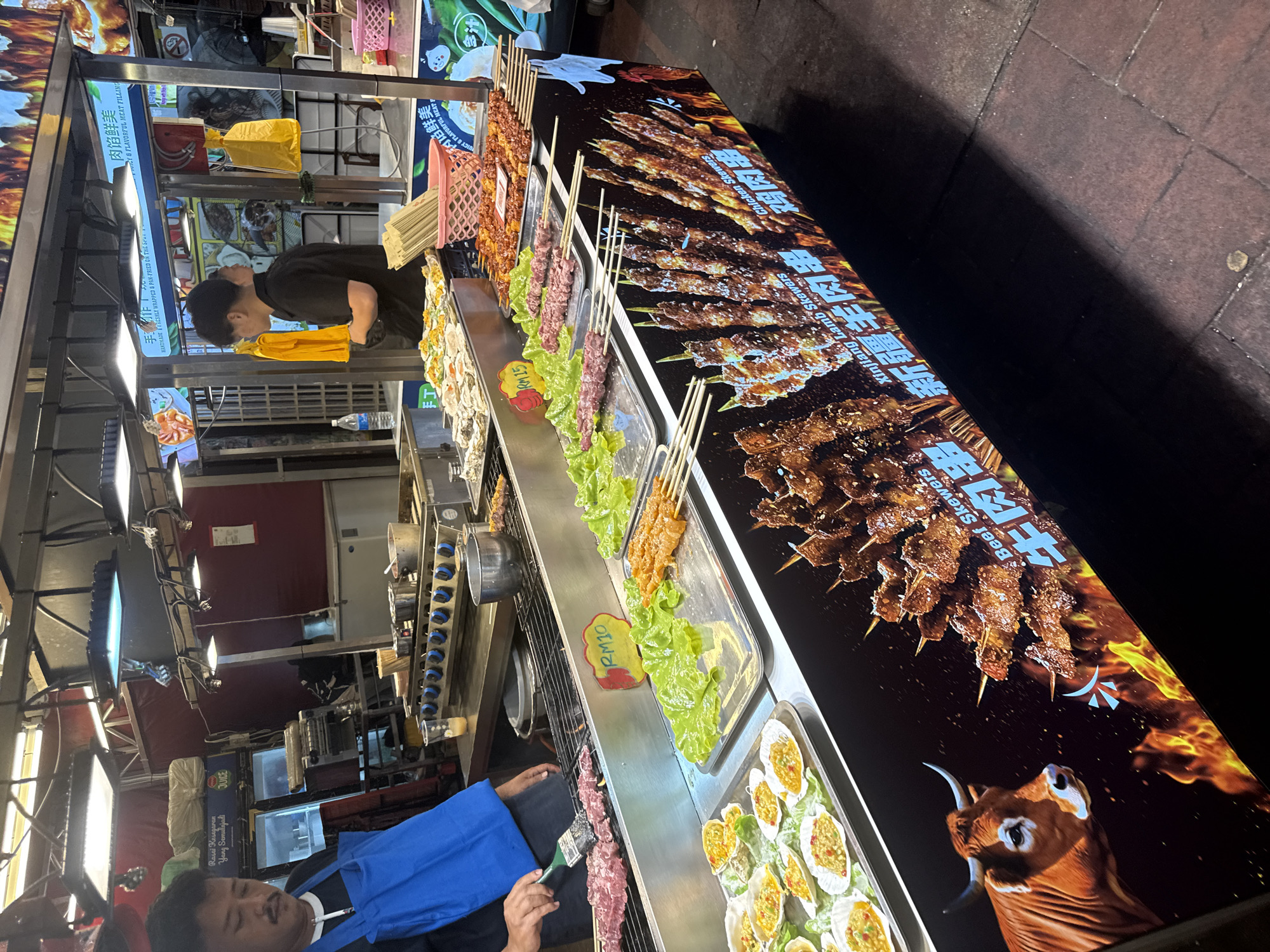 This media file captures a vibrant street food stall scene, likely at night, in Kuala Lumpur, Malaysia. The main subjects are two male vendors, various food items, and the stall's setup.
One vendor, wearing a blue apron, is actively preparing food, brushing a sauce onto raw skewers arranged on a metal tray. The other vendor, dressed in a black shirt, stands further back, seemingly managing the stall or preparing other items.
The stall prominently displays a variety of food, predominantly different types of skewers, along with what appear to be grilled oysters in shells and some form of meat pie or dumpling. The skewers include raw meat (possibly lamb and chicken) on lettuce beds, and cooked, seasoned skewers displayed on a large, illuminated sign that functions as a menu. The grilled oysters are arranged neatly in a tray, topped with a yellow, savory filling.
Notable details include the time of day, which is clearly night due to the artificial overhead lighting casting bright illumination on the food and stall, against a dark background. Pricing is visible on small yellow signs: "RM10" and "RM15", indicating Malaysian Ringgit, consistent with the provided location context.
Visible text on the stall and signs includes:
- "Rasai Kesegaran Yang Semulajadi" (Malay for "Taste the Natural Freshness")
- "手作" (Chinese for "Handmade")
- "HANDMADE FRESHLY WRAPPED & PAN-FRIED ON THE SPU"
- "肉馅鲜美" (Chinese for "Juicy & Flavorful Meat Filling")
- "JUICY & FLAVORFUL MEAT FILLING"
- "Beef Skewers" and "牛肉串" (Chinese for Beef Skewers)
- "Xinjiang Lamb Skewers" and "新疆羊肉串" (Chinese for Xinjiang Lamb Skewers)
- "Chicken Skewers" and "鸡肉串" (Chinese for Chicken Skewers)
- A large graphic on the display panel features a bull's head and the text "Beer Skewers."
The overall atmosphere suggests a busy street food environment, characterized by delicious aromas and a diverse offering of grilled and fried dishes.