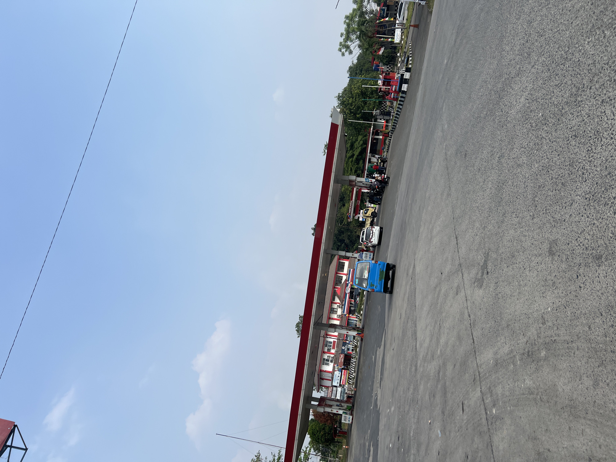 An outdoor, daytime scene at a Pertamina gas station in Gunung Putri, Indonesia, shows a paved area with several vehicles and a building. A blue, three-wheeled vehicle is positioned centrally, facing away from the camera. To the left, under a red and white covered awning of the gas station, are several motorcycles and individuals. The gas station building features red and white trim and multiple air conditioning units. Further to the right, more vehicles, including a yellow truck and white cars, are visible, as are other gas pumps. The environment includes trees and a clear blue sky with scattered clouds. A black wire is visible stretching diagonally across the upper portion of the image.