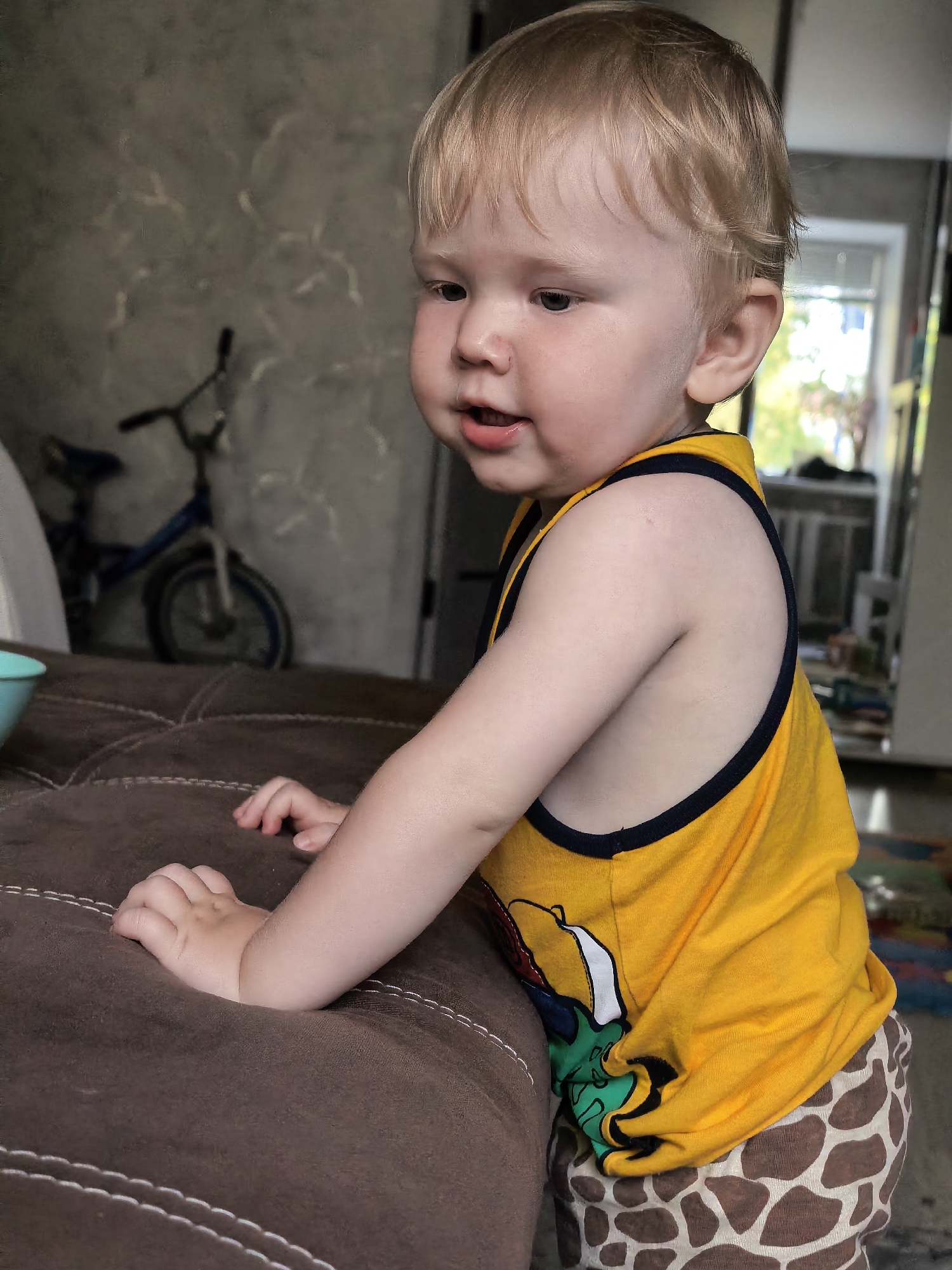 A young child with blond hair and light skin is leaning on a brown upholstered surface, possibly a couch. The child is wearing a yellow tank top with a dark trim and patterned shorts with a giraffe print. The background shows a wall with a textured pattern, a blue bicycle, and a window with greenery visible outside. The overall scene appears to be indoors, in a residential setting. The image does not contain any specific geographical markers to indicate its location within the city of Sterlitamak, Russia.