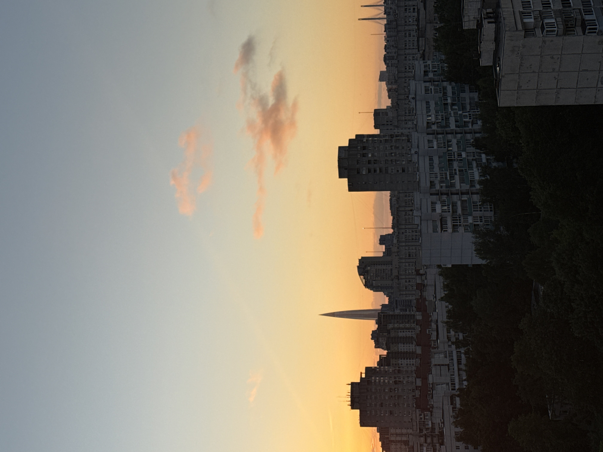 The image depicts a cityscape of Saint Petersburg, Russia, at sunset. A tall, slender, needle-like skyscraper dominates the skyline, with a city stretching out below it. The sky is a gradient of warm orange and soft blue hues, with scattered pink clouds adding color to the scene. In the foreground, a dense canopy of dark green trees obscures the lower levels of buildings. The buildings themselves appear to be a mix of older, blocky apartment buildings and more modern structures. The overall impression is one of urban sprawl under a dramatic twilight sky.