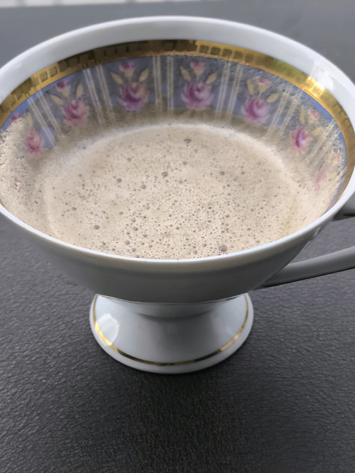 A close-up, overhead shot shows a white ceramic teacup with a gold rim and a decorative band featuring a blue pattern with pink floral accents, filled with a frothy, light brown beverage. The cup rests on a darker, textured surface, possibly a table. The background is blurred, suggesting an outdoor setting. The image is devoid of any discernible Lithuanian landmarks or elements that would place it specifically in Dercekliai, Lithuania.