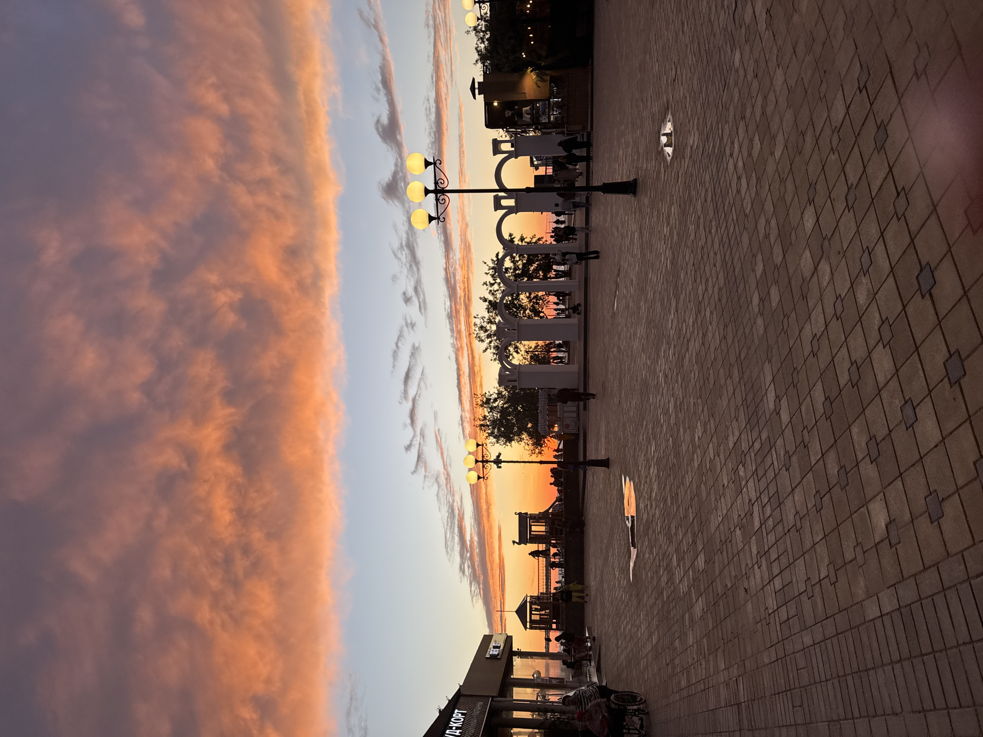 This image depicts a serene sunset scene along a paved promenade in Anapa, Russia. The sky, especially on the left, is ablaze with dramatic, elongated clouds, painted in warm hues of orange, pink, and soft purple, contrasting beautifully with the clearer blue sky above.
The foreground and midground are occupied by a wide, patterned paved walkway or square, reflecting the soft, fading light. Several ornate streetlights, featuring multiple glowing spherical lamps, punctuate the promenade, casting pools of light. Further down the path, a series of white, arch-shaped structures, possibly pergolas, create an interesting architectural line, with some trees and foliage visible between them.
The setting sun, a bright orange disc, is visible low on the horizon, casting a warm, intense glow and silhouetting figures and structures. Numerous people are visible as dark silhouettes along the promenade, appearing to walk, gather, or simply enjoy the beautiful evening. The activity suggests a leisurely atmosphere, common in a resort town during sunset.
In the bottom left corner, part of a building is visible, bearing the Cyrillic text "АУД-КОРТ" on its sign. A partial view of a bicycle and a wheelchair can also be seen near this building. The overall impression is one of peaceful enjoyment during dusk, with the vibrant sky being a prominent feature.