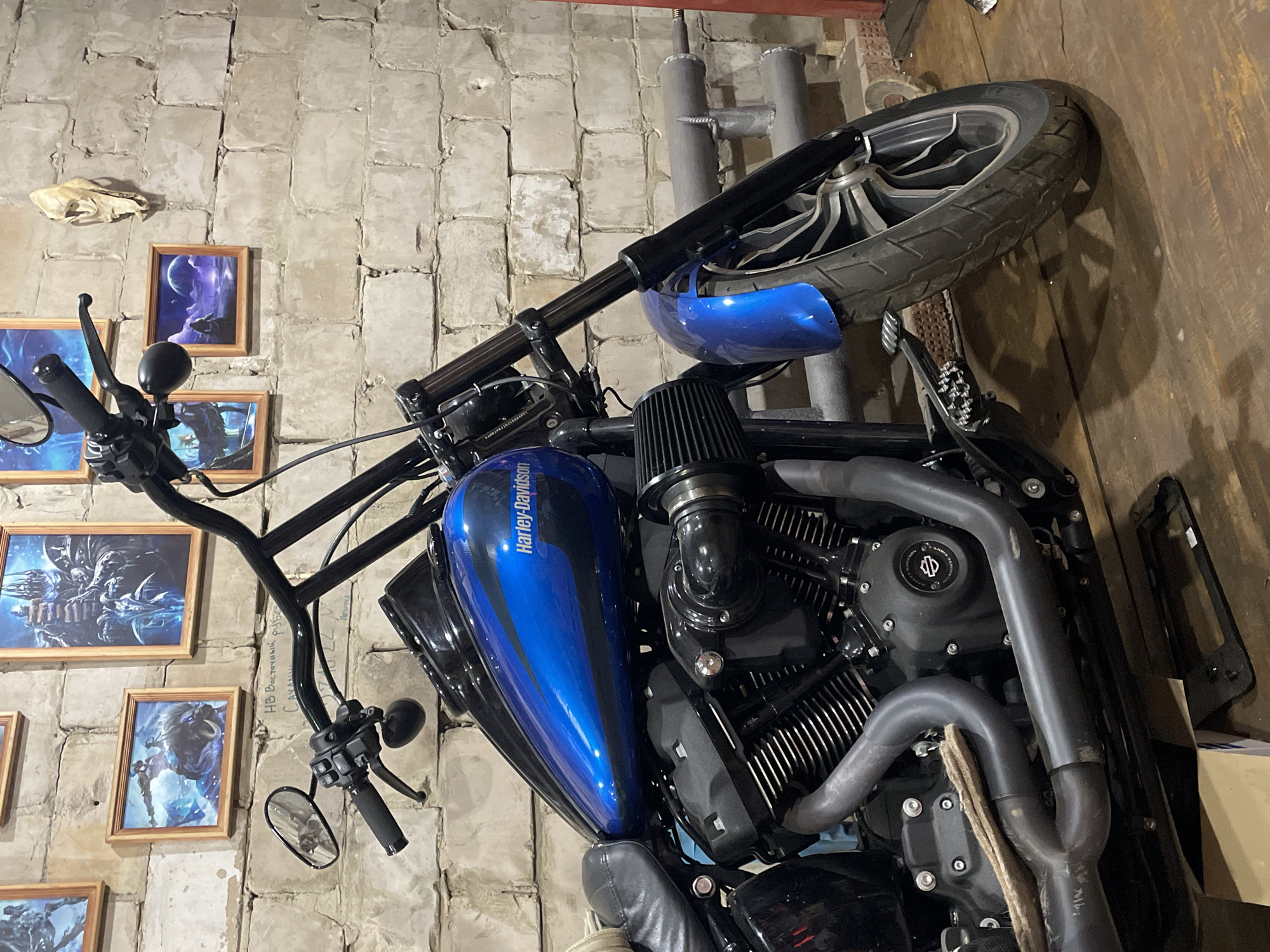 The image shows a close-up view of a Harley-Davidson motorcycle inside a garage or workshop, positioned at an angle. The motorcycle is predominantly black with a blue gas tank bearing the "Harley-Davidson" logo. The engine, exhaust pipes, handlebars, and front tire are all visible. The background is a textured brick wall with several framed pictures and what appears to be an animal skull. The scene is likely located in Oktyabr'skii, Russia. The environment suggests an informal setting, possibly a personal workspace or storage area. There are no people visible in the shot.