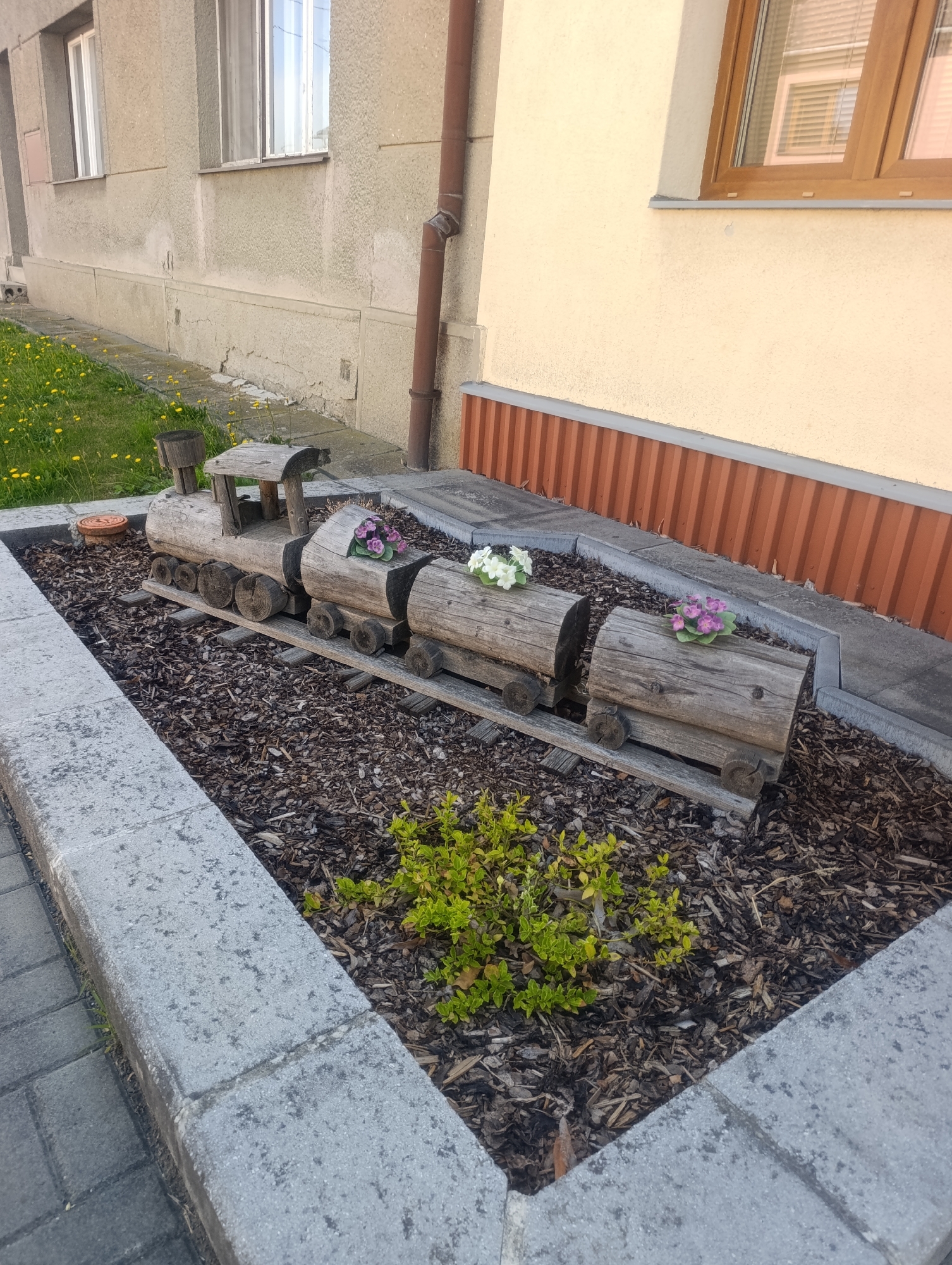 In the city of Výkleky, Czechia, a charming garden feature depicts a wooden train set placed in a flowerbed. The train, crafted from logs, consists of a locomotive and multiple cars, each adorned with colorful flowers. The flowerbed is filled with dark mulch and bordered by concrete slabs. To the left, a paved walkway and a grassy area with dandelions are visible, while a building with windows and a downspout stands behind the garden.
