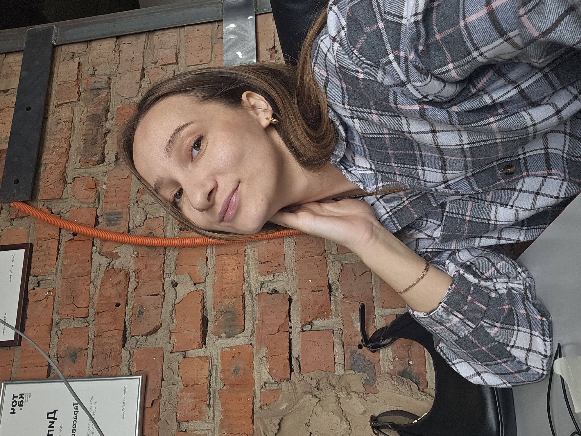 A young woman lies on a brick wall, smiling at the camera. She is wearing a gray plaid shirt and a gold bracelet. There are framed papers on the wall behind her with Russian text; "Kg POT" is visible on one frame. An orange conduit also runs along the wall. The location is identified by the OCR text on the papers in the city of Chaikovsky, Russia. A black chair is visible behind her.