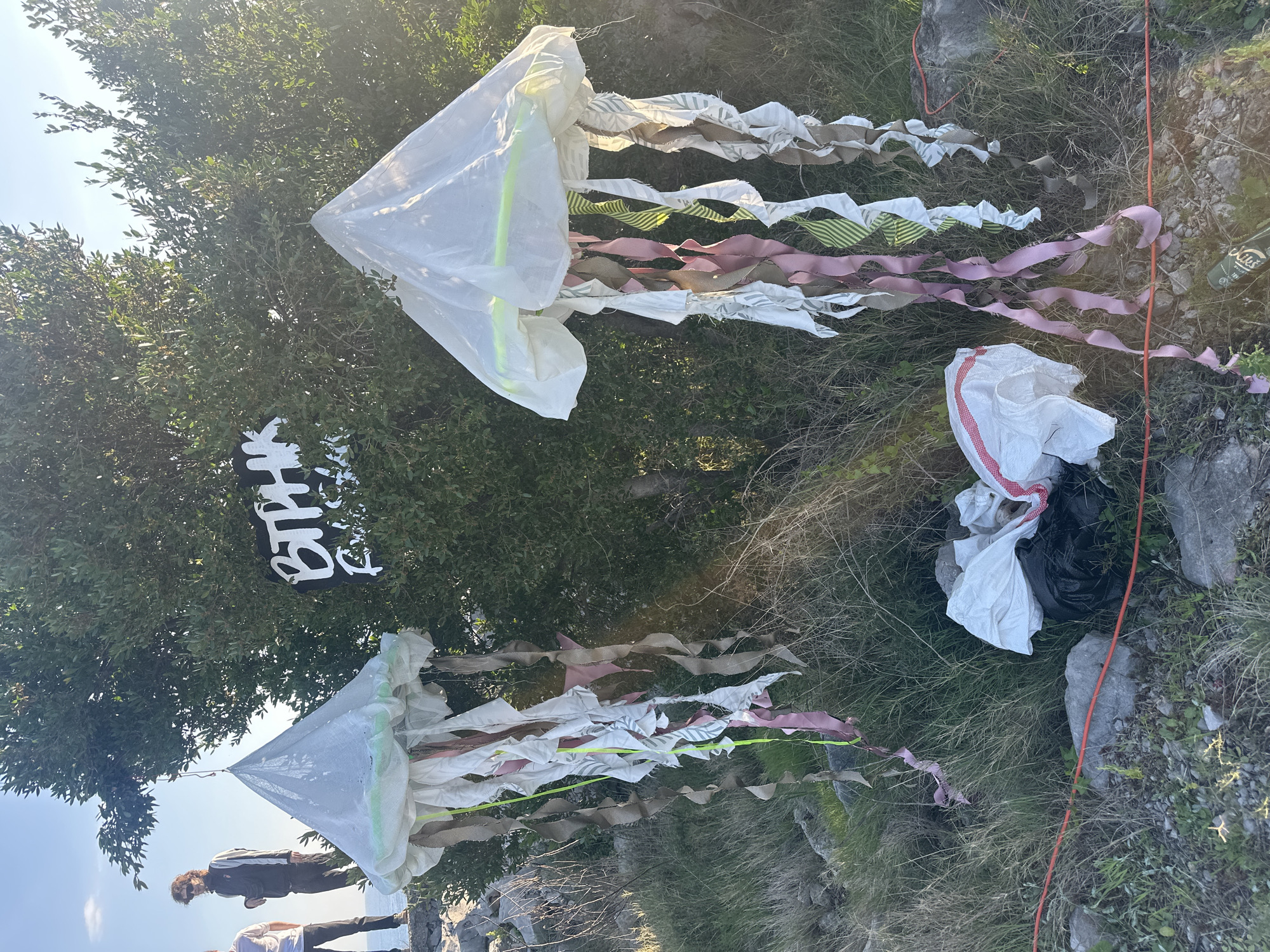 The image shows an outdoor scene in what appears to be Glavaticici, Montenegro. Two roughly cone-shaped objects, possibly art installations or decorations, are prominent. They have white conical tops and streamers made of various materials like fabric or ribbon in colors like white, pink, green, and brown. The ground cover consists of grass, rocks, and some vegetation.
In the background, a dark sign with white writing that appears to say "BTHYK" is visible. There are also what appear to be discarded white and black bags. Part of a group of people are seen along with a few rocks. The lighting suggests it is daytime and the sky is visible in the corner.