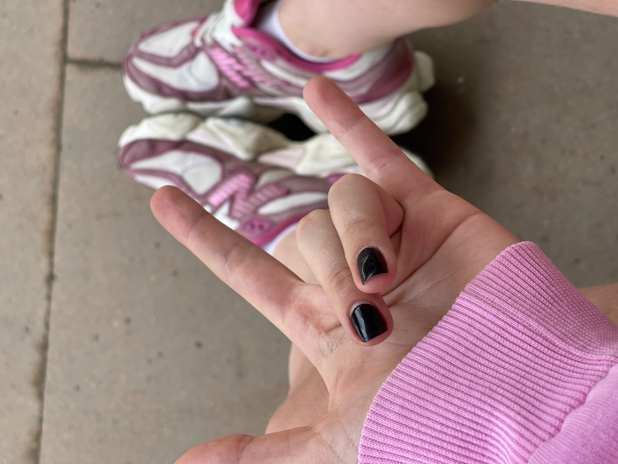 The image shows a hand making the "rock on" gesture with the index and pinky fingers extended, and the middle and ring fingers folded down, nails painted black. The hand is wearing a pink long-sleeved shirt, and in the background, there are pink and white sneakers on a concrete surface. There is no information available in the image to identify the location.