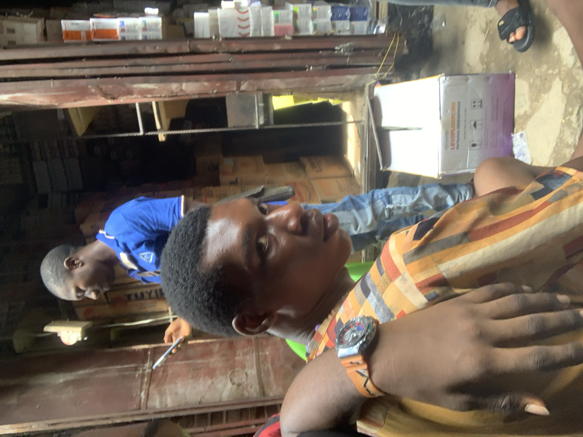 The image shows two people inside what appears to be a pharmacy or a store selling pharmaceutical products. The location is possibly in Fegge, Nigeria.
In the foreground, a person with short, dark hair is wearing a patterned shirt and a watch on their left wrist. Their hand is prominently displayed, close to the camera. In the background, another person is wearing a blue shirt and appears to be using a smartphone.
Behind the people, there are shelves stocked with various medications and pharmaceutical products. A white box with the text "LEVOFLOXACIN" is visible. The environment is somewhat cluttered, suggesting a busy commercial area. Outside the building, part of a leg wearing a sandal can be seen.