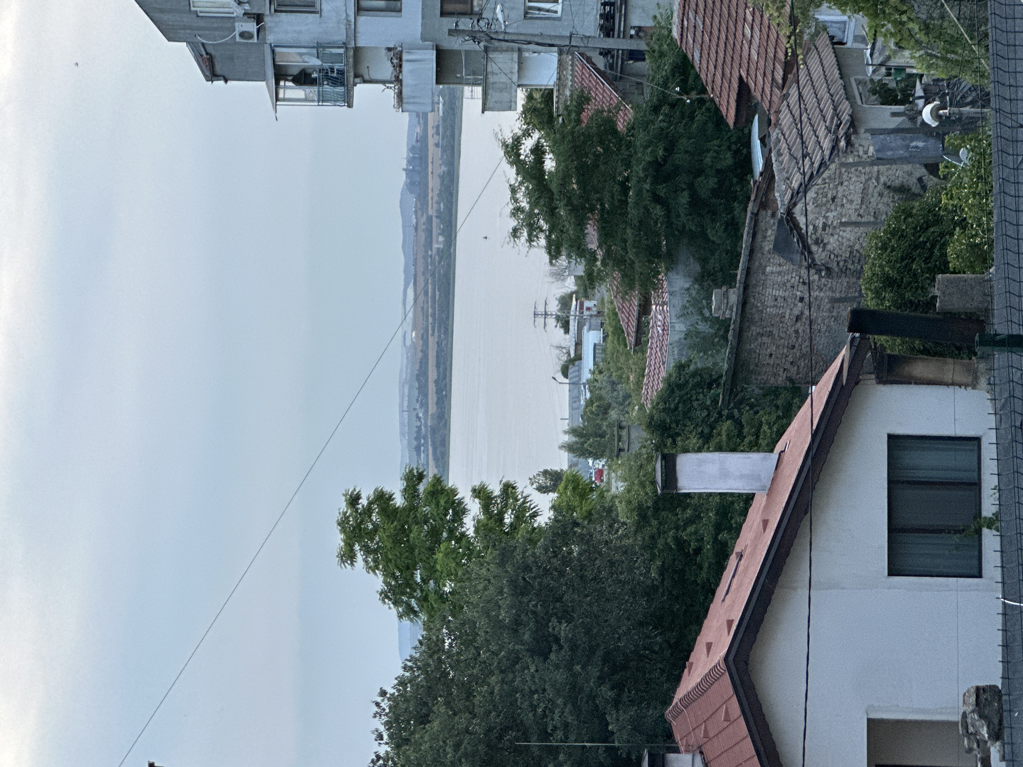 The image depicts a view of the Varna Lake, Bulgaria, from a residential area. In the foreground, a white house with a red tiled roof and a prominent chimney is visible. To the right, a multi-story grey building with balconies stands out, with a person on one of the upper balconies. Green trees are abundant throughout the scene, framing the lake and the buildings. The lake itself is calm and reflects the overcast sky. In the distance, across the lake, there are industrial structures and hilly terrain. The overall impression is of a partly urban, partly natural landscape under a cloudy sky.