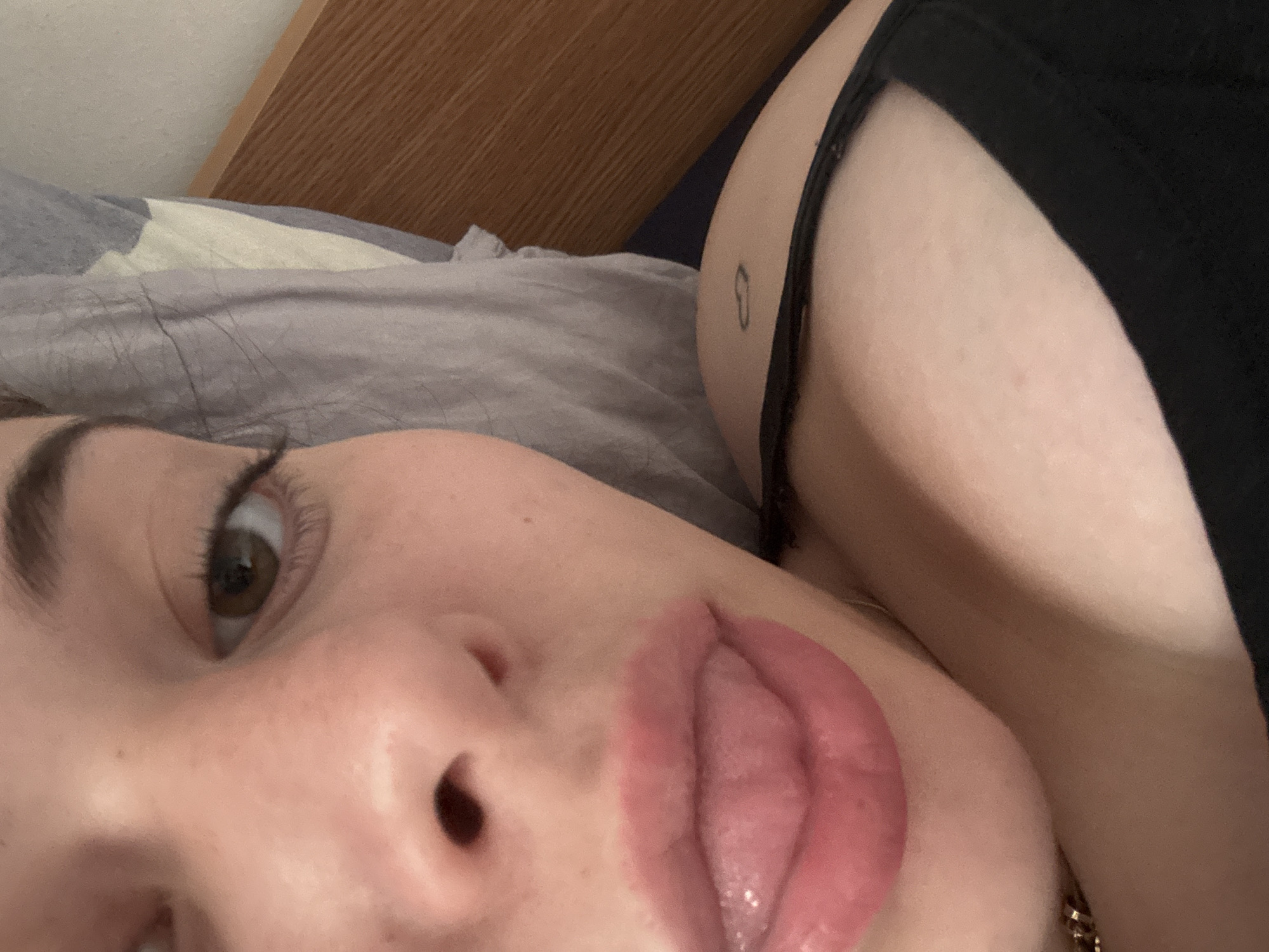 A close-up, angled selfie depicts a fair-skinned person with light brown eyes and dark eyebrows lying on what appears to be a bed with a light gray patterned sheet and a wooden headboard. The person's lips are prominent, coated in a pinkish gloss, and their tongue is extended. Visible skin displays freckles and blemishes.
Behind the person, a bare shoulder and upper back are visible, marked with a small tattoo and adorned with a black garment strap. A gold chain necklace is also partially visible around the neck.
There is no visible evidence in the image to confirm the location as Troyan, Bulgaria.