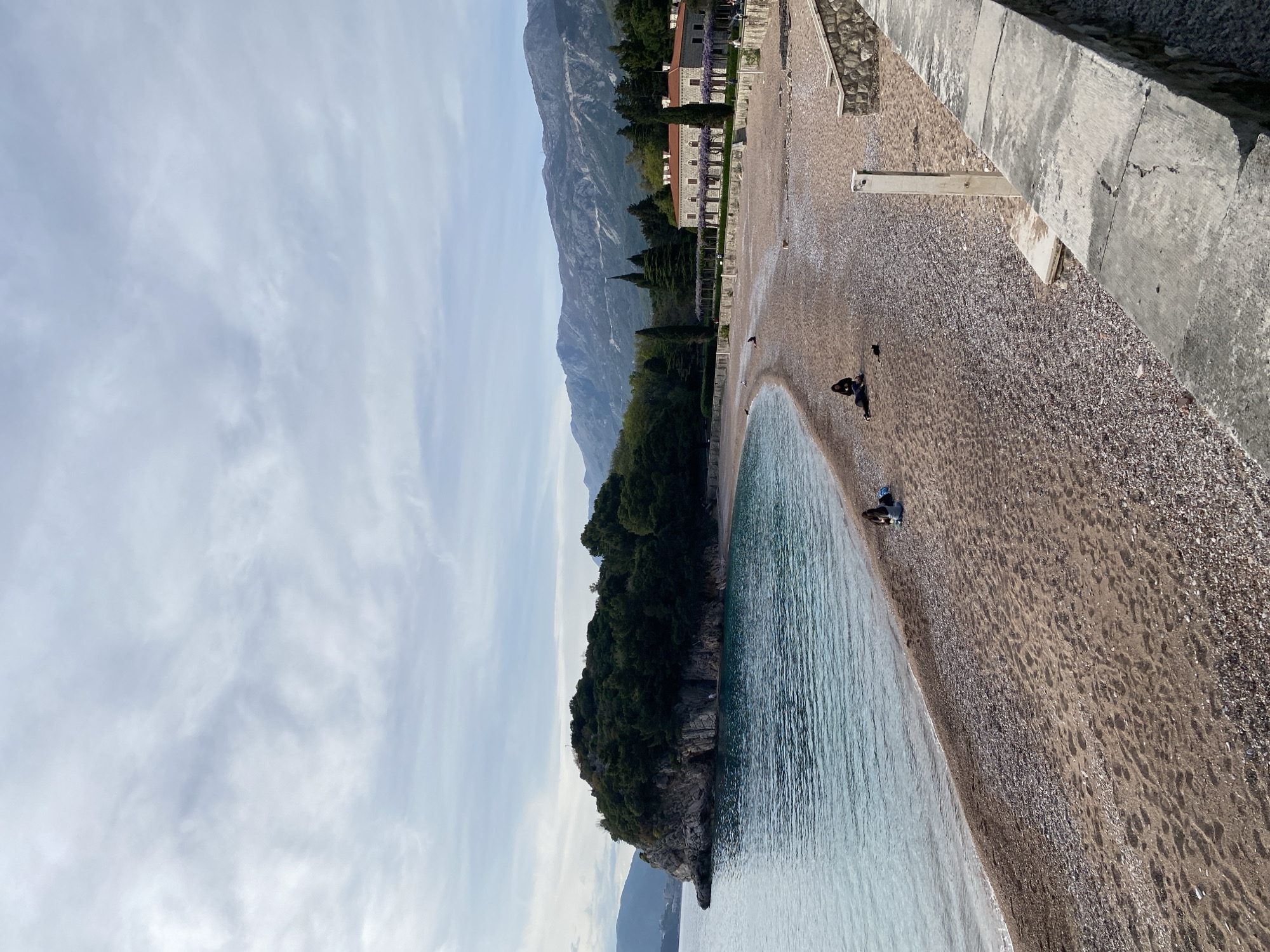 The image depicts a beach in Sveti Stefan, Montenegro. The beach curves along the shoreline, with the sea on one side and a rocky cliff covered in trees on the other. In the distance, mountains rise up against a blue sky with scattered clouds. Along the beach, several people are visible, some appear to be walking or relaxing. The beach surface varies from sand to small pebbles and rocks. The beach is bordered by a stone wall, behind which buildings are visible.