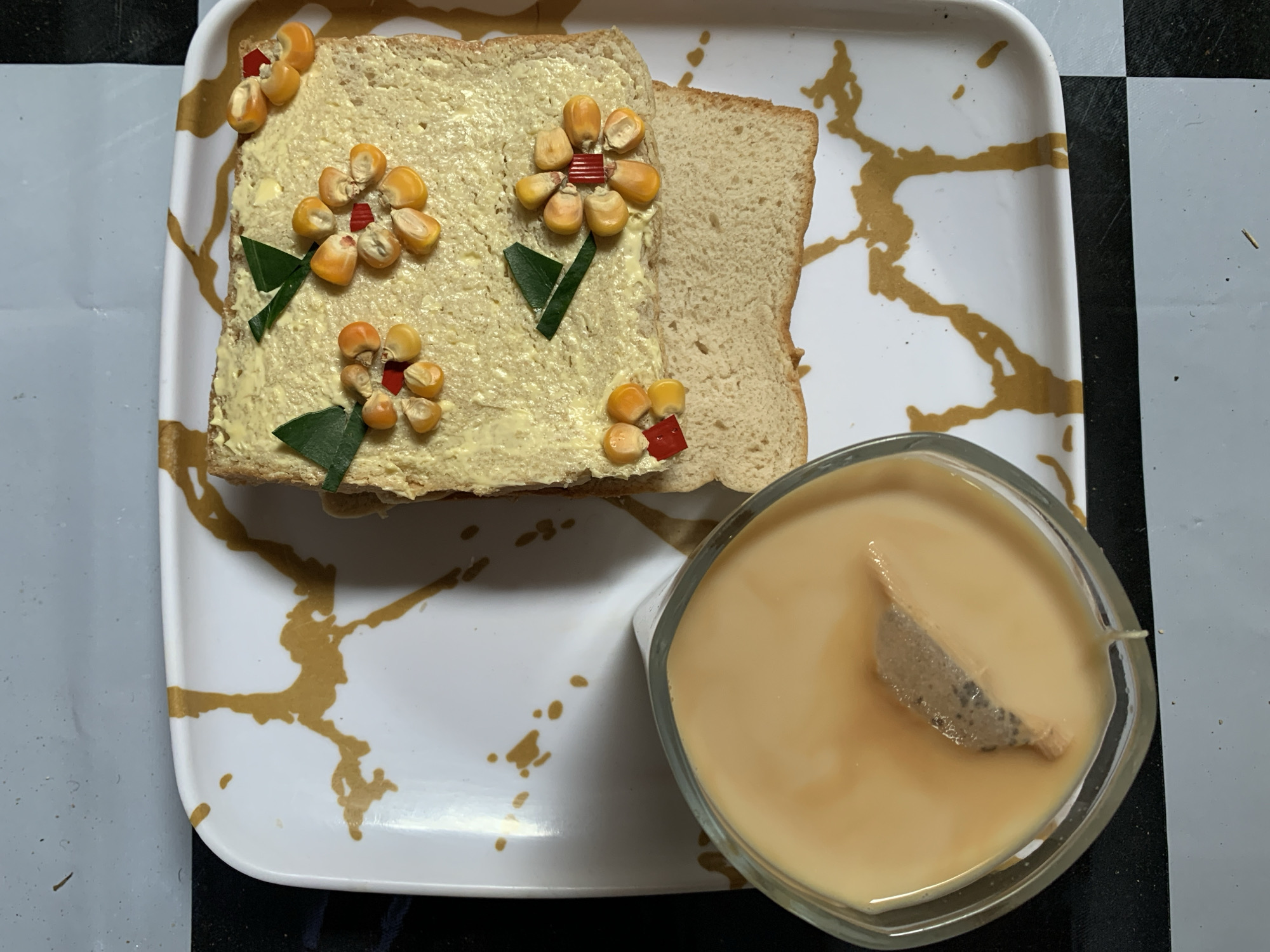 On a white plate with gold-colored abstract decorations, there is a sandwich made of two slices of bread. One slice is spread with a yellow substance and decorated with corn kernels arranged like flowers, with small green leaves and red pieces as accents. Next to the sandwich is a clear glass containing a light-brown liquid, in which a tea bag is steeping. The plate rests on a surface with a black and white checkered pattern and gray sections. The location is Rafin Par, Nigeria.
