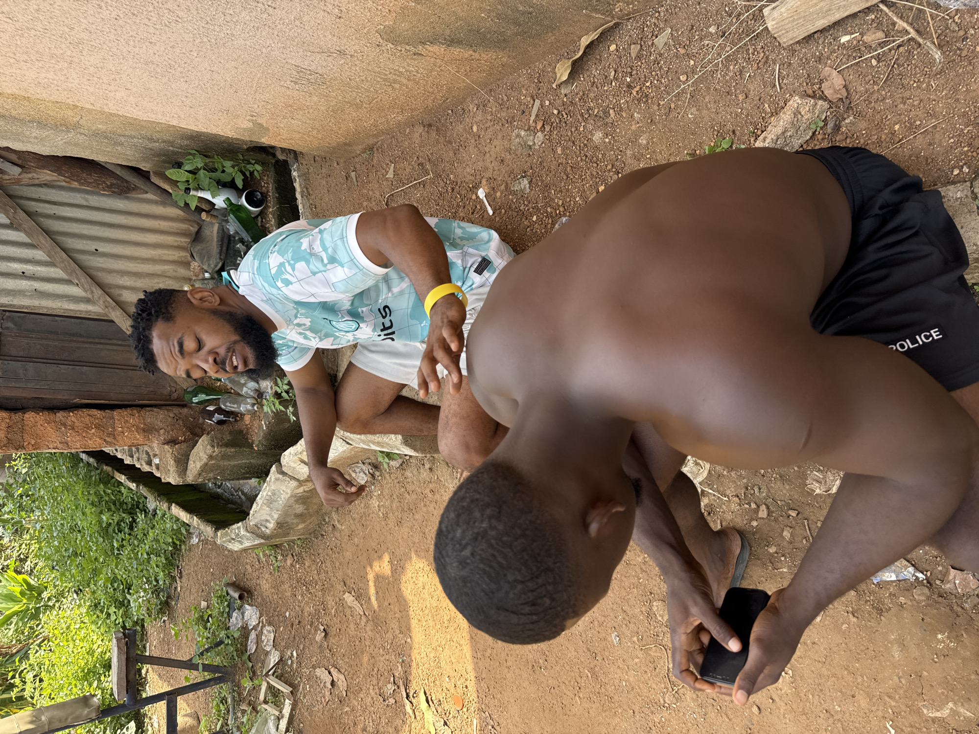The image depicts an outdoor scene in Akwanga, Nigeria. Two Black males are present. The foreground subject, viewed from the back, is crouched down, looking at a black smartphone in his hands. He has short, dark hair and is wearing black shorts with "POLICE" written in white text on the left leg. The second subject is seated, facing left, with his eyes closed and mouth slightly open. He has dark skin, a beard, and short, curly hair. He is wearing a light blue and white patterned t-shirt and white shorts, with a bright yellow wristband on his left wrist. The environment is rural, with dirt ground, scattered debris, and overgrown vegetation. A concrete drainage channel runs along the side of a building constructed of mud bricks and corrugated metal. Several empty glass bottles are visible near the building. A metal chair with a wooden seat is in the background to the left. The lighting suggests it is daytime.