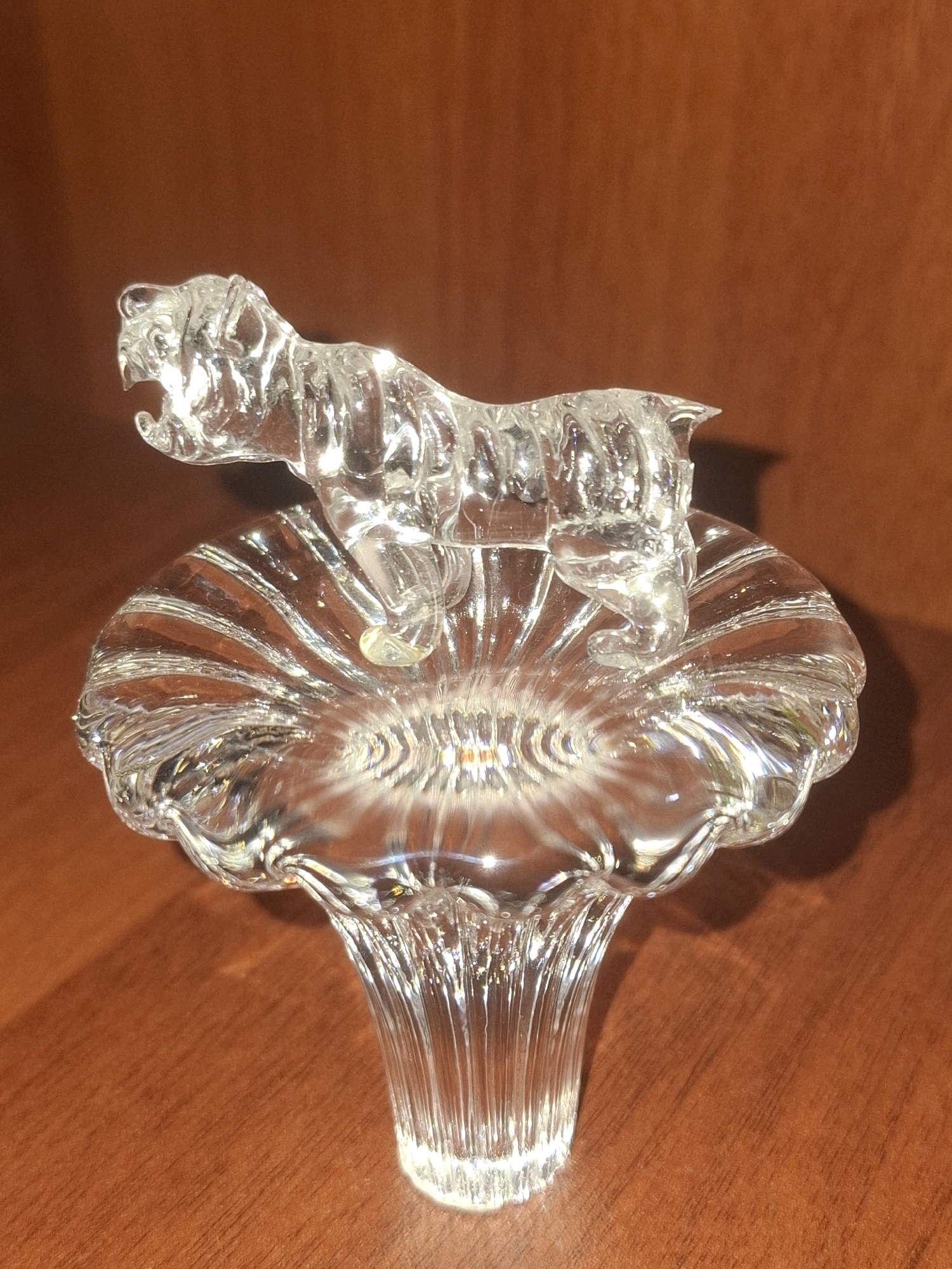 A clear glass figurine of a tiger stands on top of a clear glass decorative piece with a fluted top and a ribbed stem, resembling a mushroom. The tiger is in mid-stride with its mouth open. The objects are placed on a polished wooden surface. The background is a warm brown, out-of-focus wall. The image is an indoor still life. There is no information in the image that indicates the location of Almaty, Kazakhstan.