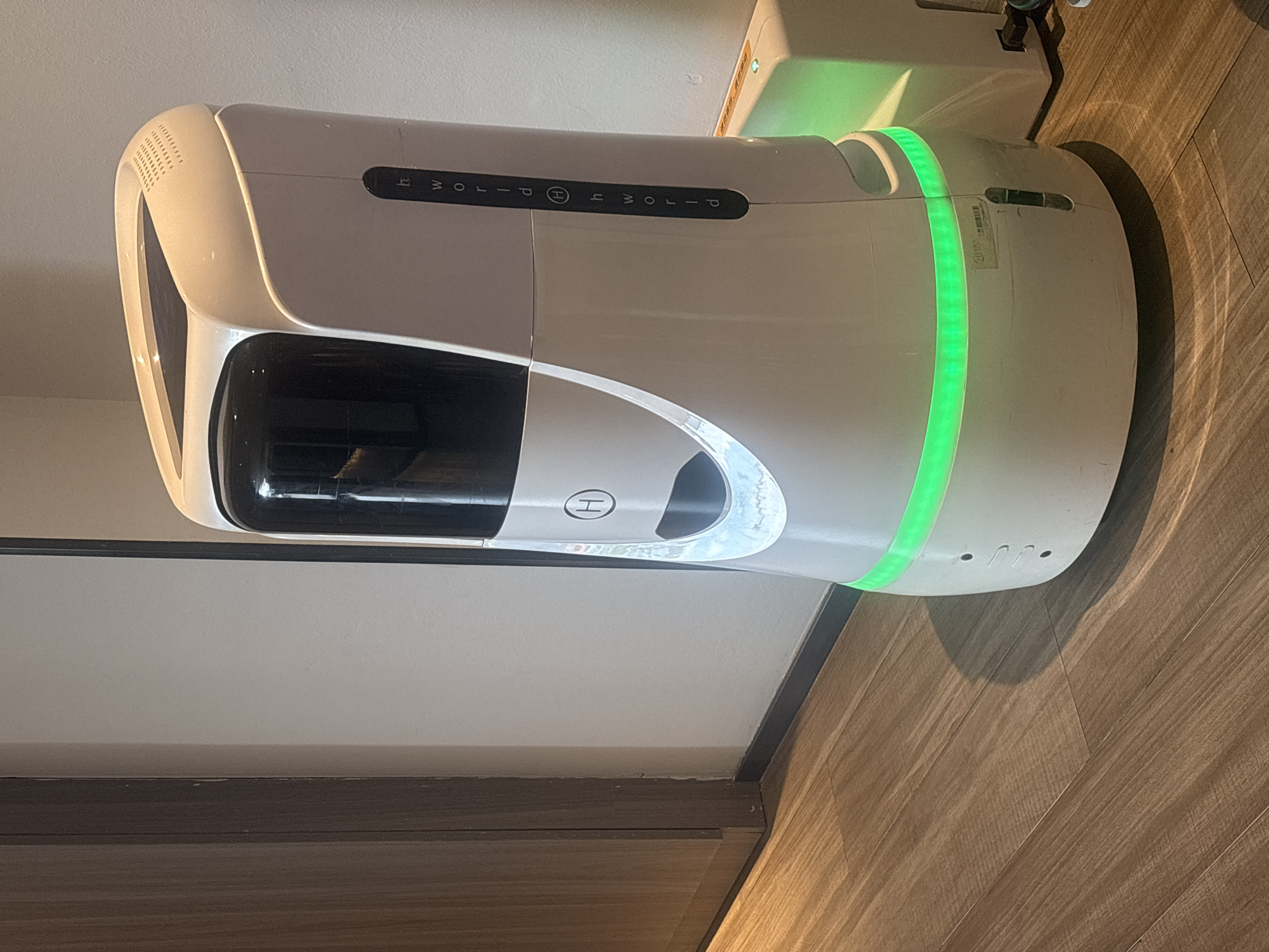 The image shows a white delivery robot with a black screen and a green LED strip around its base. The robot is standing on a wooden floor next to a white wall and a closed wooden door. The robot has the letters "hworld" printed vertically on its side, with a circular "H" logo in the middle. A small rectangular indicator light is visible on a white box to the right of the robot, which is glowing green. This scene is likely located within the Suzhou Industrial Park, China, given the context of the provided information. There are no people visible in the image.