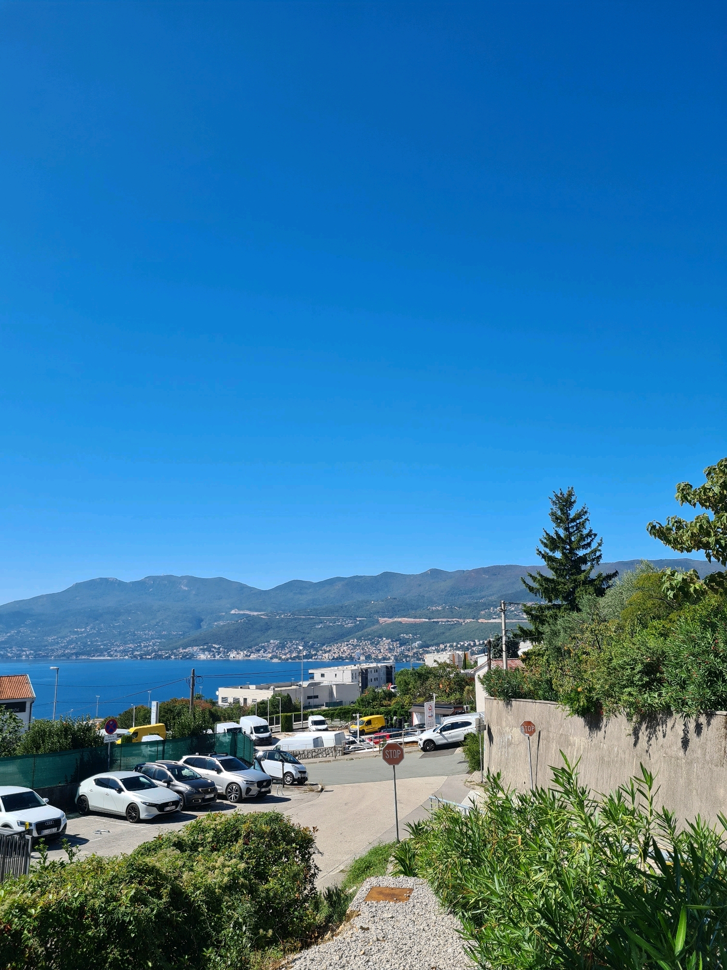 The image shows a clear blue sky over a coastal city. In the foreground, there is a parking lot with several cars, including white, grey, and silver sedans and SUVs. A white van is also visible. Lush green bushes and trees line the parking lot and a concrete wall. In the middle ground, there is a road with more vehicles, including a yellow van. Buildings, some white and modern, are scattered throughout the scene. Beyond the city, a body of water, likely the Adriatic Sea, is visible, bordered by a mountainous coastline. The mountains are covered in vegetation and dotted with small towns or villages. The overall impression is a bright, sunny day in a Mediterranean coastal area. The location is identified as Grad Rijeka, Croatia, which is a major port city in the northern Adriatic.