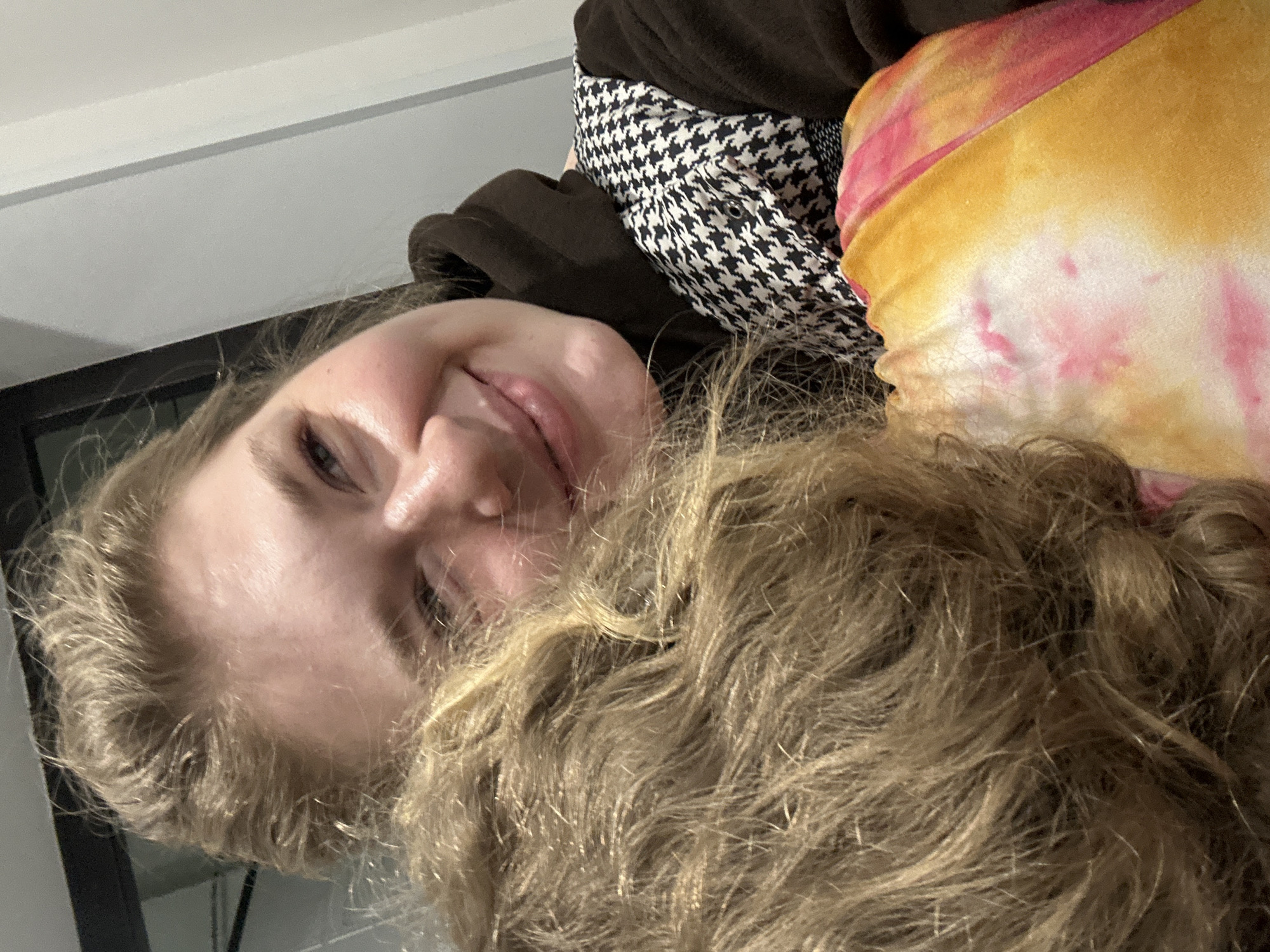This image depicts an indoor scene in Kazan, Russia, featuring two individuals in a close-up, affectionate interaction. An adult female with fair skin and light-colored hair pulled back is visible from the chest up on the right side of the frame, smiling and looking towards the viewer. She wears a dark brown hooded sweatshirt underneath a black and white houndstooth pattern vest or sleeveless top. Resting against her right shoulder, facing away from the viewer, is a second individual, whose head is largely obscured, revealing only the back and top of their wavy, light-blonde hair. This individual is wearing a garment with a multi-colored tie-dye pattern, primarily pink, orange, and white, visible on the left shoulder. The background consists of a plain, light-colored wall on the right and a dark, indistinct vertical element on the upper left. The scene is well-lit, highlighting the subjects.