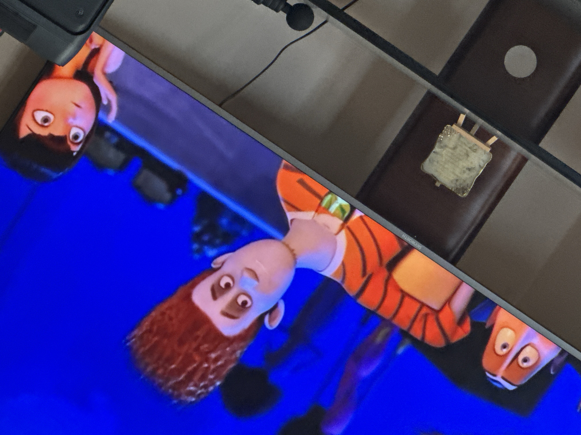 A television screen displays an animated scene featuring cartoon characters with exaggerated features against a blue background. A computer speaker is visible in the top left corner, and a power cable and a piece of furniture are positioned in the upper center. A desk with a small object on it appears in the top right. The scene is lit by artificial lighting. The city of Yerevan and Armenia cannot be determined from the image content.