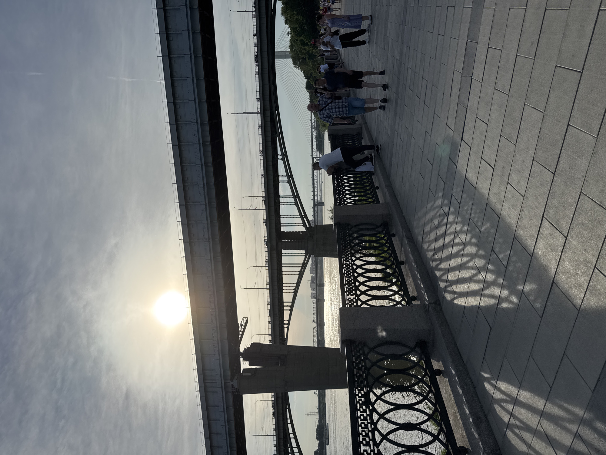 The image captures a wide shot of the embankment along the Ob River in Novosibirsk, Russia. In the foreground, a paved walkway is bordered by a decorative black metal fence with stone posts. The sun is visible in the sky, casting long shadows from the fence onto the walkway. Several people are walking and standing on the embankment. In the background, a series of bridges span the river, with the most prominent being a large railway bridge. The scene is illuminated by the warm light of the setting or rising sun.