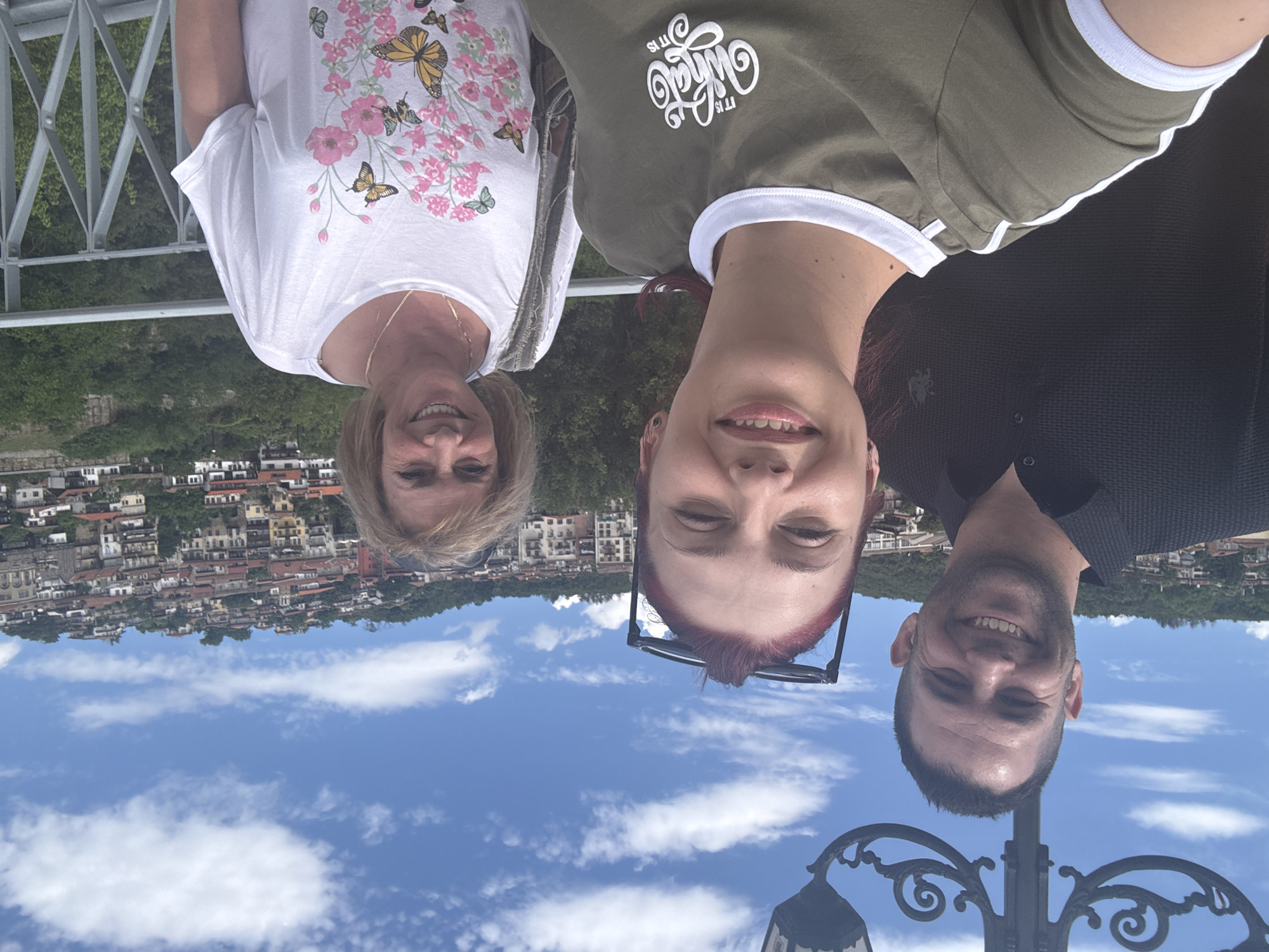 In a selfie taken on a sunny day, three people are positioned in front of a cityscape. A man on the left, wearing a black collared shirt, smiles broadly. Next to him, a young woman with red hair, wearing a green t-shirt with white trim and the text "it is what it is" printed on it, also smiles. On the right, a woman with blonde hair, wearing a white t-shirt adorned with a floral and butterfly print, smiles warmly. Behind them, the picturesque city of Veliko Tarnovo, Bulgaria, unfolds on a hillside, with its characteristic red-tiled roofs and traditional architecture cascading down the slopes. The sky is blue with scattered white clouds, and a decorative lamppost is visible in the upper left corner. The group is standing on a bridge or railing, overlooking the city.