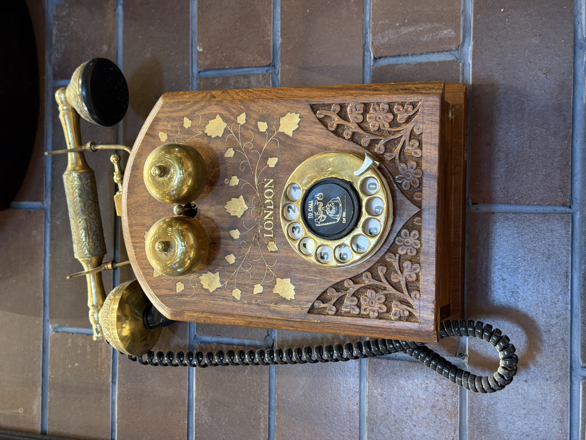 The image displays an antique-style wall-mounted telephone, set against a background of reddish-brown rectangular tiles with grey grout lines. The telephone's main unit is a wooden plaque, richly decorated with carved and gilded elements. On the right side of the wooden plaque, there are deep, intricate floral carvings. The left side is adorned with gold leaf patterns of vines and leaves, and the word "LONDON" is inscribed vertically in capital letters within this gold design.
A brass candlestick-style handset is attached to the left side of the wooden unit. It features a patterned brass handle, a black earpiece at the top, and a brass mouthpiece. Two small, circular brass bells are mounted above the handset's resting place on the wooden plaque. On the right side of the wooden unit, there's a brass rotary dial with numbers 0 through 9 clearly visible. The center of the dial contains text: "TO CALL" with an arrow pointing to a small telephone icon, and "Ext No............". A coiled black cord extends from the bottom right of the wooden unit, suggesting its connection to an unseen receiver or power source.
The phone appears to be a decorative item, showcasing vintage craftsmanship. No activity is taking place; the telephone is static and mounted on the wall. The setting is a tiled surface, possibly indoors in Gangamuthanahalli, India. The time of day and weather are not discernible.