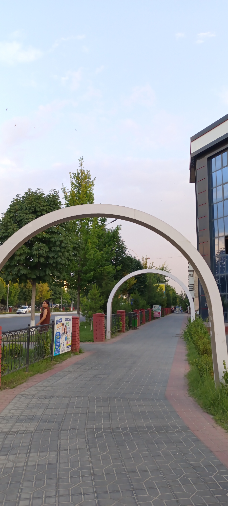 The image shows a paved walkway in Fergana City, Uzbekistan, lined with trees and decorative arches. The walkway is made of gray pavers arranged in a geometric pattern, bordered by red bricks. On the left side of the walkway, a black metal fence separates it from a road where a white car is visible. A woman in a brown outfit stands by the fence, looking away from the camera.
Further down the walkway, several posters are attached to brick pillars. One poster, in particular, is visible and features text that appears to be in English and possibly another language, with images of people. The walkway is framed by white, curved arches, creating a tunnel-like effect. Beyond the arches, more trees and greenery are visible, with a modern building with dark windows on the right side of the frame.
In the distance, several people are walking along the pathway. The sky is clear with a few faint clouds, suggesting it is either early morning or late afternoon. The overall atmosphere of the scene is peaceful and urban, with a hint of natural beauty provided by the trees and the decorative elements of the walkway. The lighting suggests a mild weather condition.