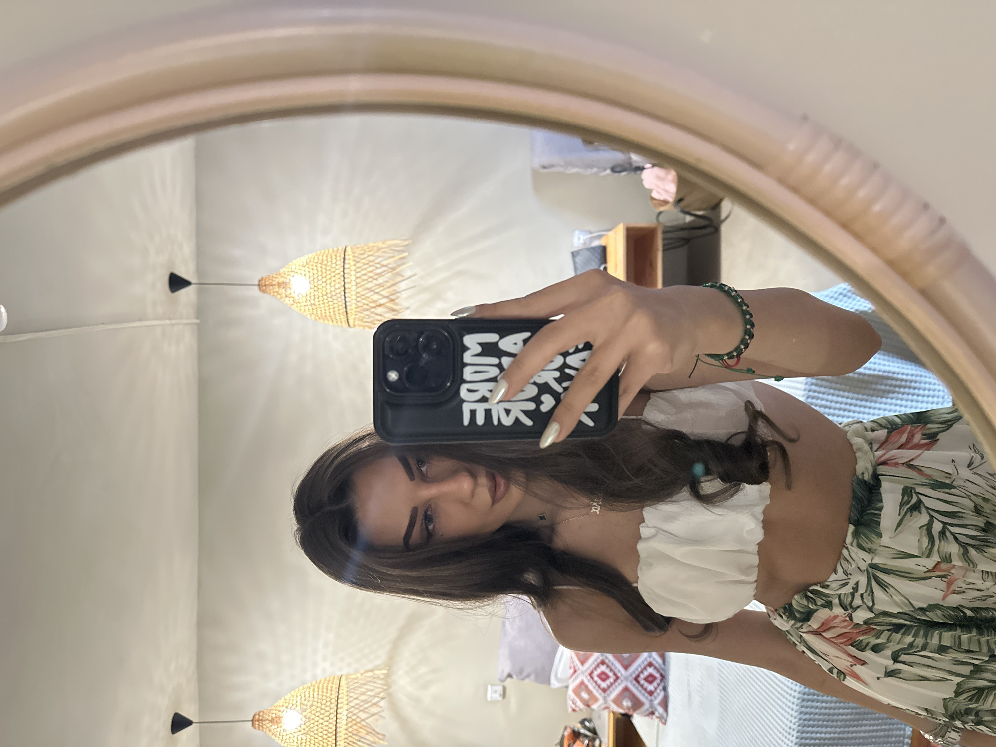A young woman with long, dark hair takes a mirror selfie in what appears to be a hotel room. She is wearing a white crop top and a flowing skirt with a tropical print. In her right hand, she holds a black smartphone with a case that reads "MORE THAN ENOUGH". Her left arm, adorned with a green beaded bracelet, is visible to the left of the phone. The background of the room shows a bed with a blue and white patterned cover, a patterned pillow, and two woven pendant lights hanging from the ceiling. The mirror has a light-colored, textured frame. The overall impression is of a relaxed, holiday setting. The location is identified as Nesebar, Bulgaria.