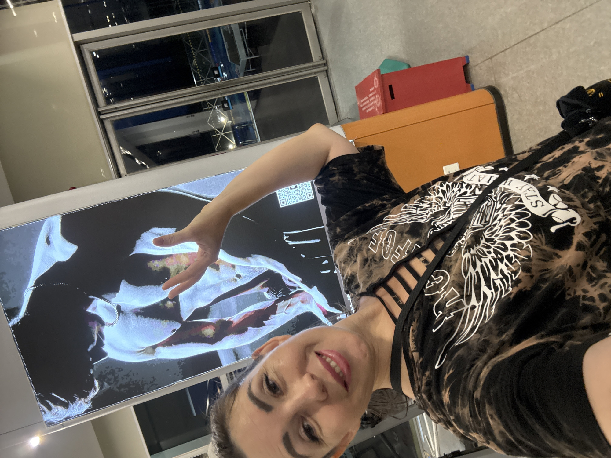 A woman with dark hair pulled back, wearing a tie-dye t-shirt with a cut-out neckline and a white graphic design of wings, smiles at the camera. She is standing in front of a large vertical display featuring a muscular figure with a blue and red hue, and a QR code. To the right of the display, large glass doors reveal a night scene with lights and structures outside. In the foreground, a red bin and an orange structure are visible. The woman is positioned in what appears to be a public indoor space, possibly a transportation hub or entertainment venue. There is no identifiable information to place this image in Huizhou, China.