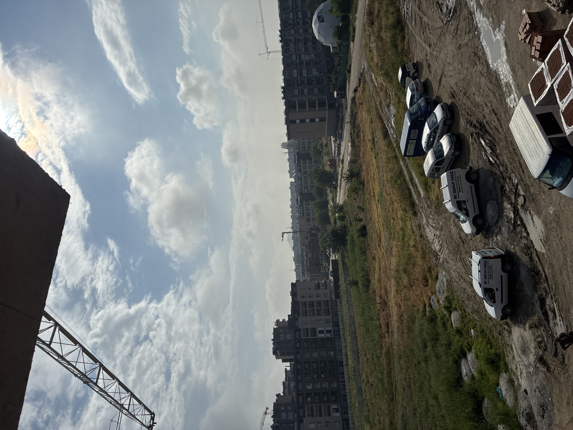 The image presents an elevated view of an urban landscape in Plovdiv, Bulgaria, during daytime under a cloudy sky. The sun, visible in the upper left, casts a subtle iridescent halo or sun dog effect in the clouds.
In the middle ground, numerous multi-story buildings, characteristic of residential or commercial structures, extend across the scene. A distinctive white geodesic dome building and at least two prominent construction cranes signify ongoing development or an industrial area.
The foreground features a wide expanse of unpaved, muddy ground, deeply rutted with vehicle tracks and containing small puddles. Areas of dry grass and weeds are also visible. Several vehicles, mainly white and one blue utility vans, are parked on this uneven terrain. On the far right, stacks of red bricks or tiles are neatly arranged on pallets next to another white van. A blurry, dark figure, possibly a person, is partially visible in the extreme lower right corner near the stacked materials.
On the sides of multiple white vans, the text "МГ ПАУЪР ЕООД" and the phone number "0884 37 77 87" are clearly legible. The scene depicts a functional, likely construction-related, environment.