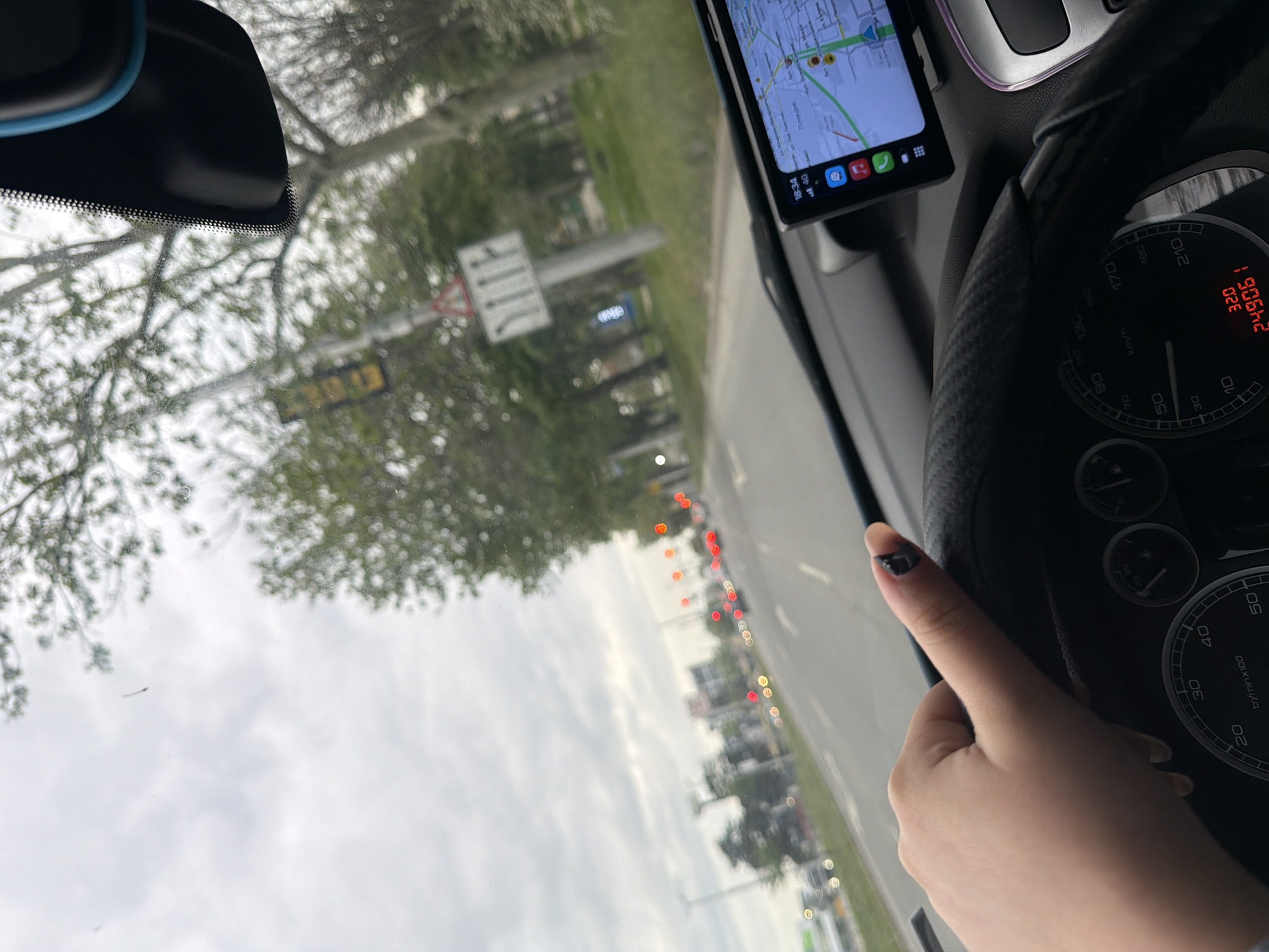 The image is taken from inside a car, likely a driver's perspective, showing the dashboard, steering wheel, and a phone displaying a map of Plovdiv, Bulgaria. The speedometer reads approximately 32 km/h and 249061. A hand is visible near the steering wheel. The car is on a road, with other cars visible ahead. The weather appears overcast. The map on the phone displays the location as Plovdiv. Road signs are visible outside the windshield.