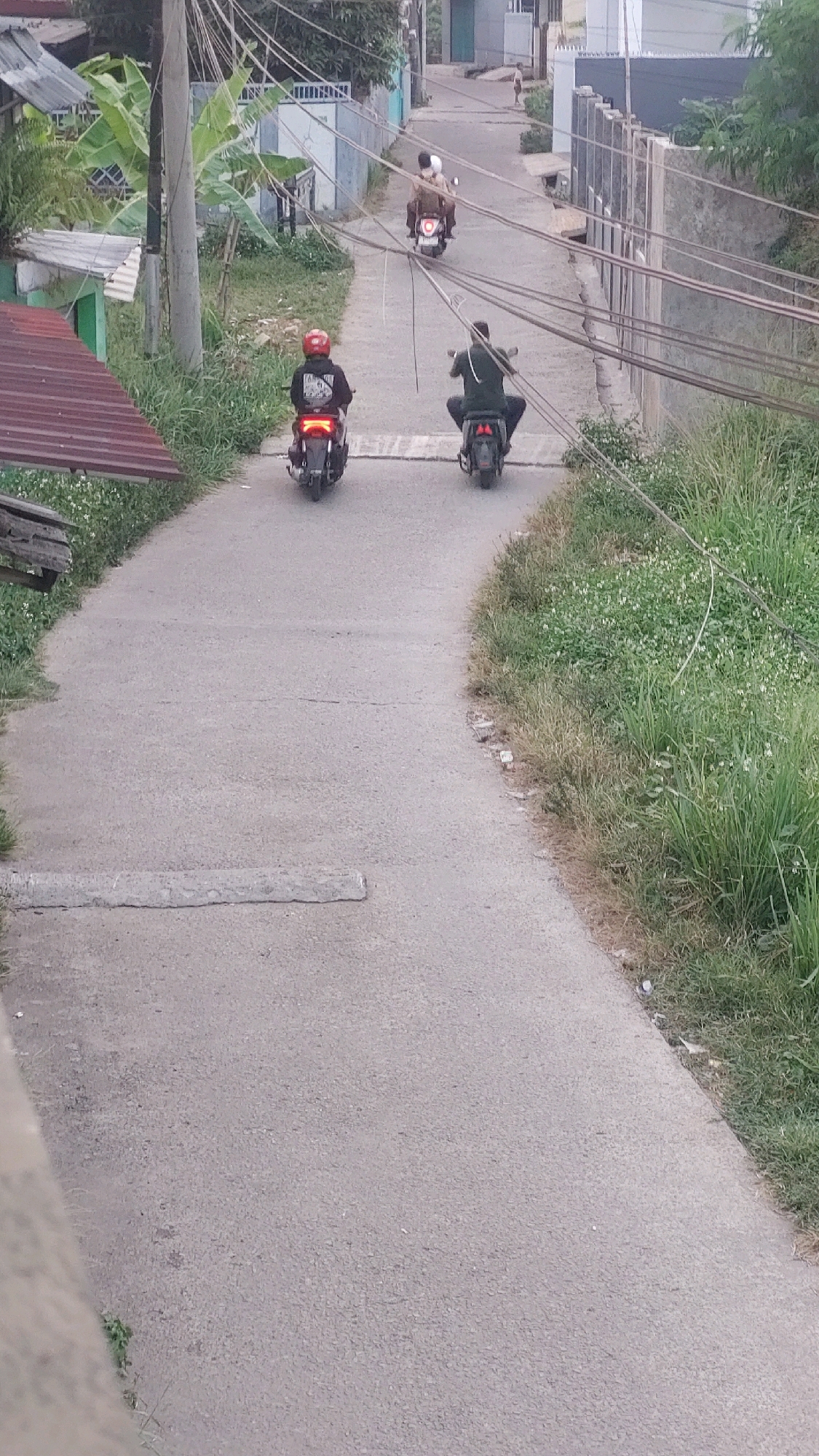 In Bojong Gede, Indonesia, on a grey, paved road, three individuals are riding motorcycles. The first motorcycle, on the left, is ridden by a person wearing a red helmet and a black jacket, facing away from the camera. The second motorcycle, further up the road and slightly to the right, carries two passengers. The driver wears a light brown top and a white head covering, while the passenger behind them is also wearing a light brown top and a white head covering. The third motorcycle, on the right, is ridden by a person wearing a dark green shirt and a dark head covering, also facing away from the camera. The road is lined with green grass and vegetation on both sides, with a row of utility poles and overhead wires running along the right side of the image. In the background, several buildings and a small, distant figure are visible. A concrete speed bump is present on the road in the foreground.