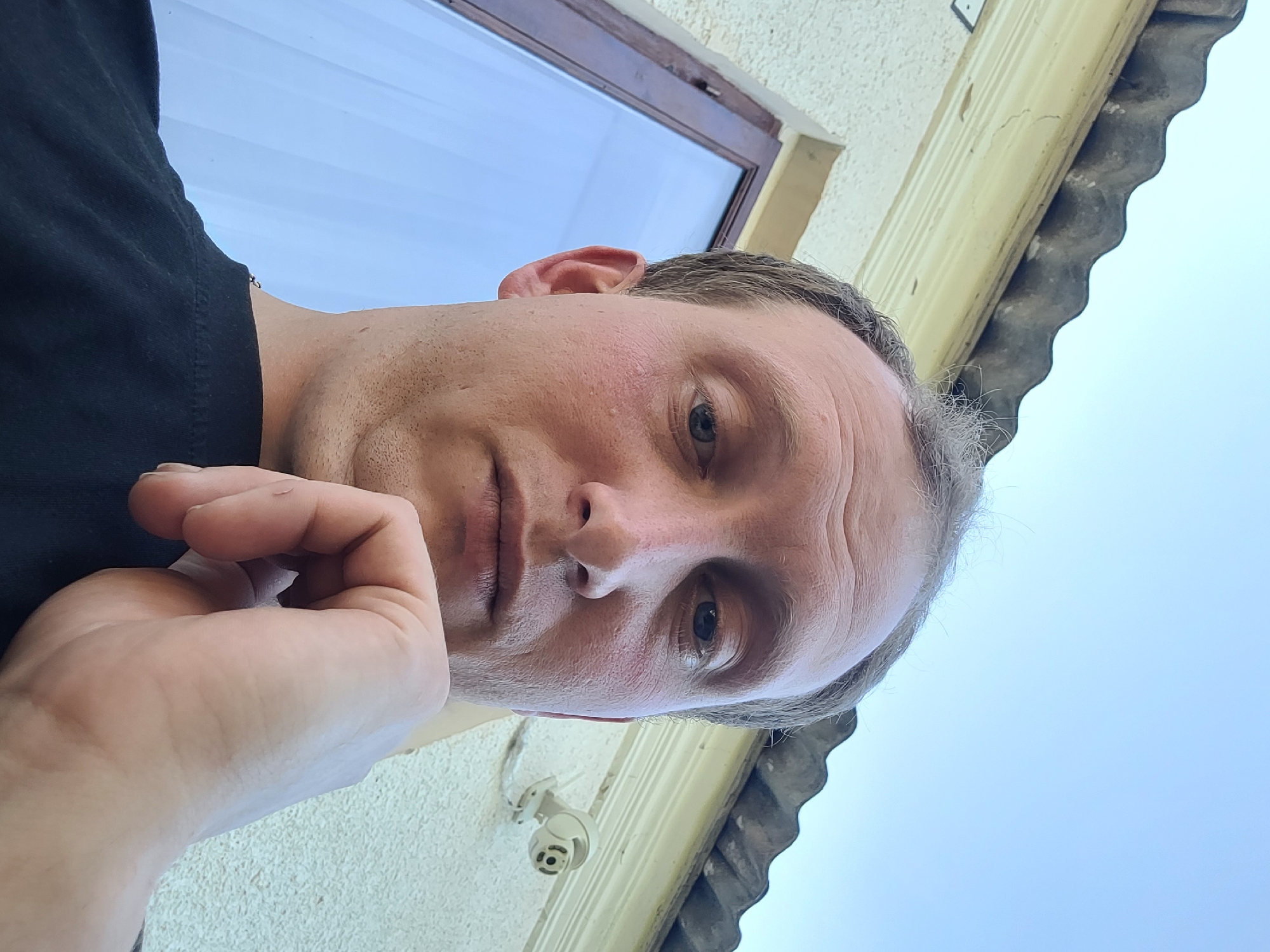 A man is captured in a low-angle, close-up selfie taken outdoors. He has fair skin, thinning light brown hair, and is resting his chin on his hand. He is wearing a black t-shirt. The background shows a cream-colored textured wall, a window with blue sky reflected in it, and a dark corrugated roofline. A white security camera is mounted on the wall to the right. The sky is clear and blue.