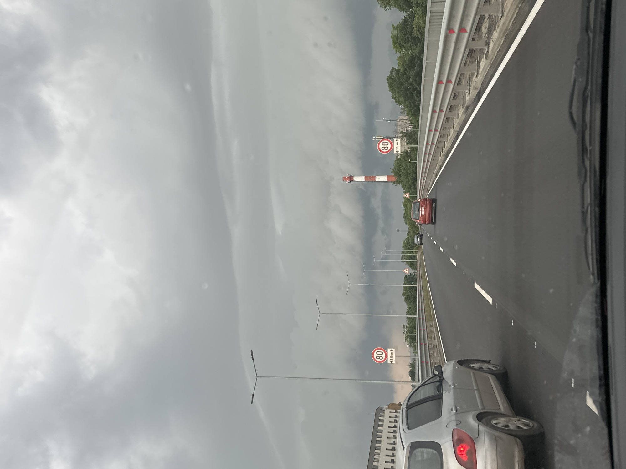 The image provides a driver's-eye view from a car on a multi-lane highway under an overcast sky. In the immediate foreground, the rear portion of a silver hatchback is visible on the right side. Ahead, a red car travels in the right lane, and a dark-colored vehicle is in the far distance in the left lane.
The highway features clear white dashed lane markings and a solid white line defining the right shoulder, which is bordered by a metal guardrail adorned with red reflectors. Modern streetlights are spaced along the left side of the road.
In the mid-ground, a tall, distinctive red and white striped structure, resembling a chimney or industrial tower, rises against the cloudy backdrop. Further left, a large, multi-story building can be seen. Green trees line sections of the highway on the right.
The weather is characterized by a heavily overcast sky filled with dense, grey clouds, indicating a dull, potentially rainy day.
Two circular speed limit signs are visible, each displaying "80" in red text on a white background with a red border, indicating an 80 km/h speed limit. Beneath one of these signs, a small white rectangular sign with an upward arrow reads "↑3.5 км↑", likely signifying a distance or stretch of road.