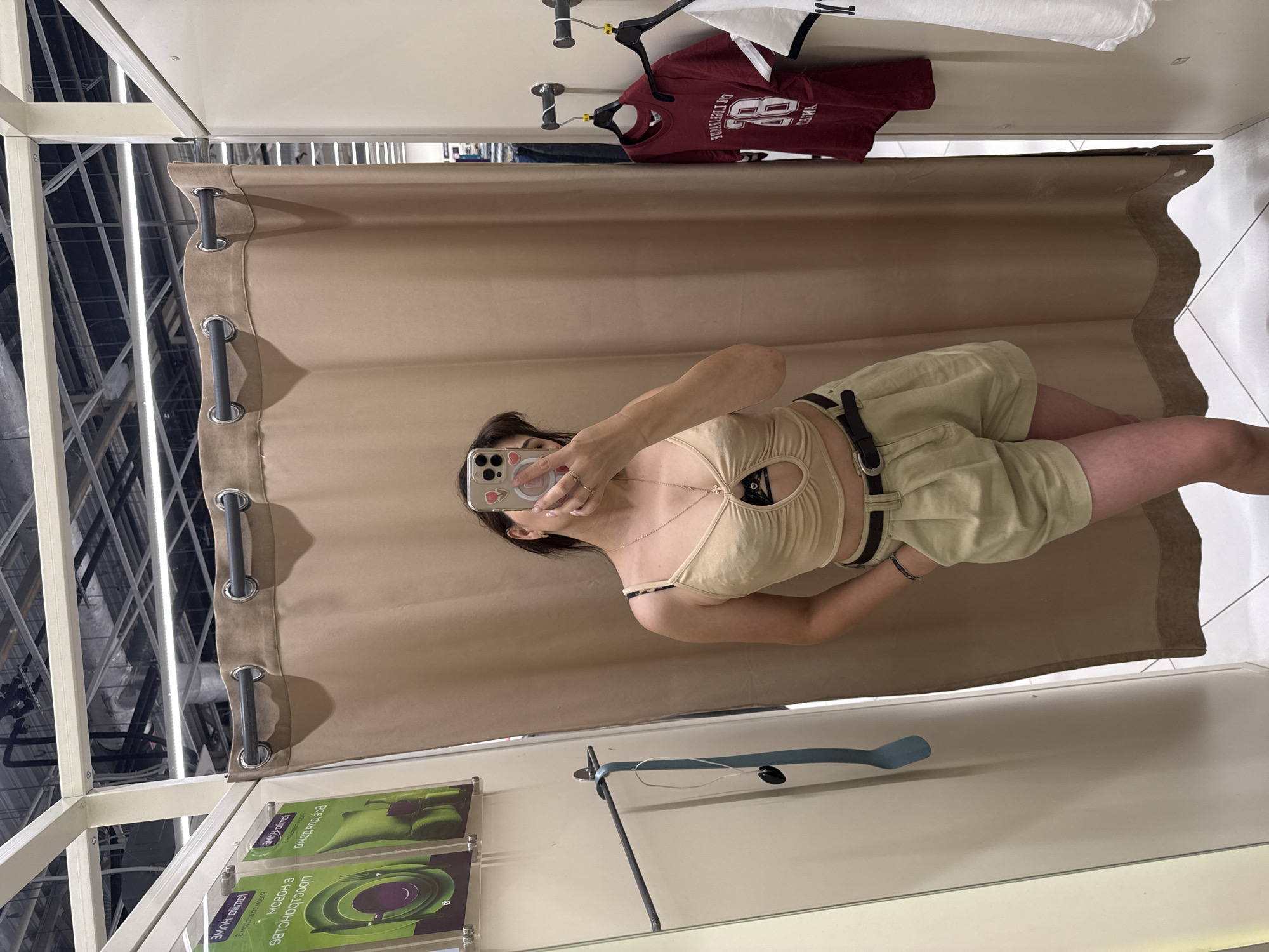 A young woman is standing in a fitting room in what appears to be a retail store. She is taking a mirror selfie with her phone. She is wearing a light beige crop top with a cut-out detail in the front, and light khaki-colored shorts with a dark belt. She is also wearing a necklace and bracelets. In the background, there are clothes hanging on racks and display boards on the wall. The fitting room has a beige curtain and mirrors. The overall environment suggests a commercial space, likely a clothing store in a city.