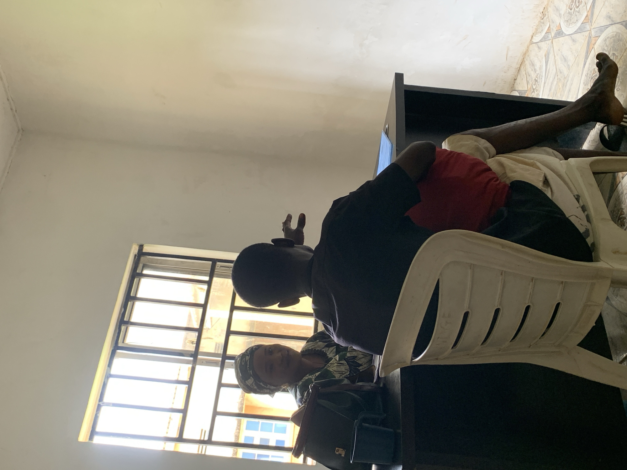 The image captures an indoor scene in Maigizo, Nigeria. A young man is seated on a white plastic chair, wearing a black shirt and red shirt, with dark shorts. His bare feet are visible, resting partially on a dark-colored cabinet or desk. Seated at the desk is a woman wearing a headscarf and a patterned top. She appears to be looking at the person behind the camera. Beside her is a dark-colored handbag. Bright sunlight is coming in through a window with metal bars, creating a bright environment. The walls are painted white, and the floor has patterned tiles.