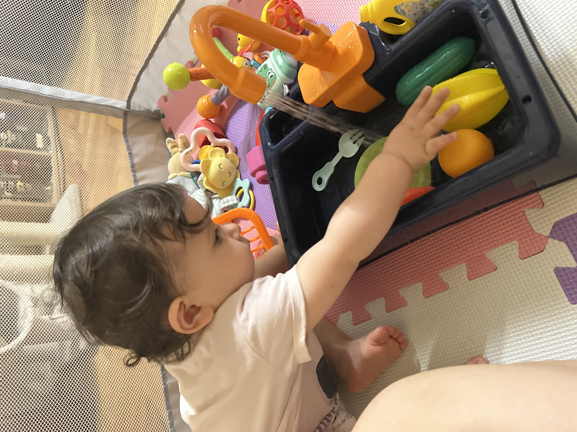 A baby is playing with a toy sink in a playpen. The baby is wearing a pink t-shirt and has dark curly hair. The toy sink is orange and has a faucet with water running. There are plastic fruits and vegetables in the sink, including an orange, a yellow starfruit, and a green cucumber. Other toys, such as a yellow plush toy with flower petals and a green ball on a stick, are visible in the background. The playpen is set up on a multi-colored foam mat. The scene appears to be indoors. The image does not provide any information about the location in the city of Sofia, Bulgaria.