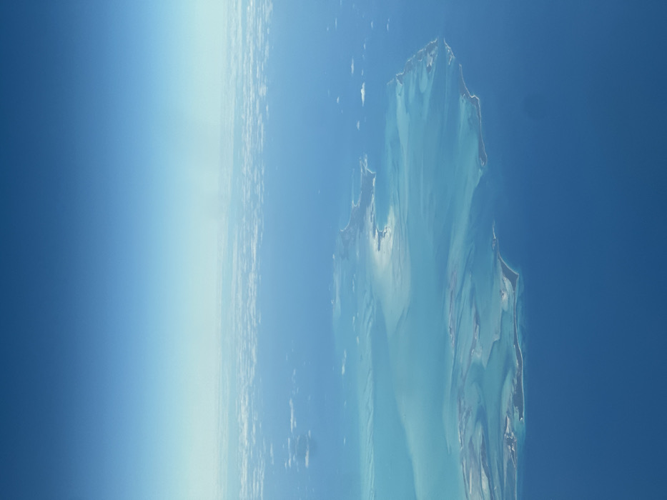 From an elevated perspective, possibly space, an island chain stretches into the deep blue sea. The turquoise waters around the islands blend with the ocean, creating varying shades of blue. On the left, a faint atmospheric glow hints at the Earth's curvature. The islands themselves appear rugged with distinct shapes and sandy shores.