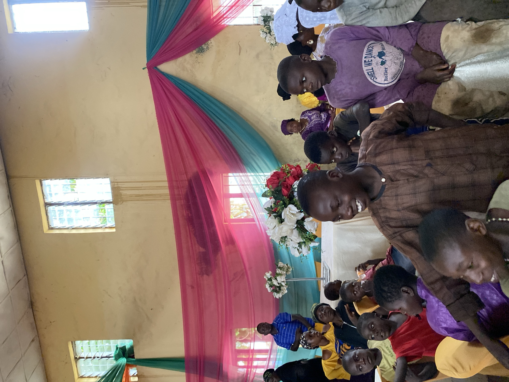 The image captures an indoor gathering, likely a ceremony or event, in Riyom, Nigeria. A group of children and adults are seated, facing forward, suggesting they are observing something happening off-camera. The room appears to be decorated with draped pink and teal fabric, creating a festive backdrop. Two windows with louvered panes are visible on the light-colored wall. In the foreground, several children are prominent, some smiling and looking towards the front. One child in the center, wearing a brown patterned shirt, is smiling broadly. To their right, a child in a purple outfit with the text "SHELL WE DANCE" is seated. In the background, a man in a blue striped shirt stands near some floral arrangements. The lighting is a mix of natural light from the windows and possibly some artificial interior lighting. The overall atmosphere seems to be one of communal observance.