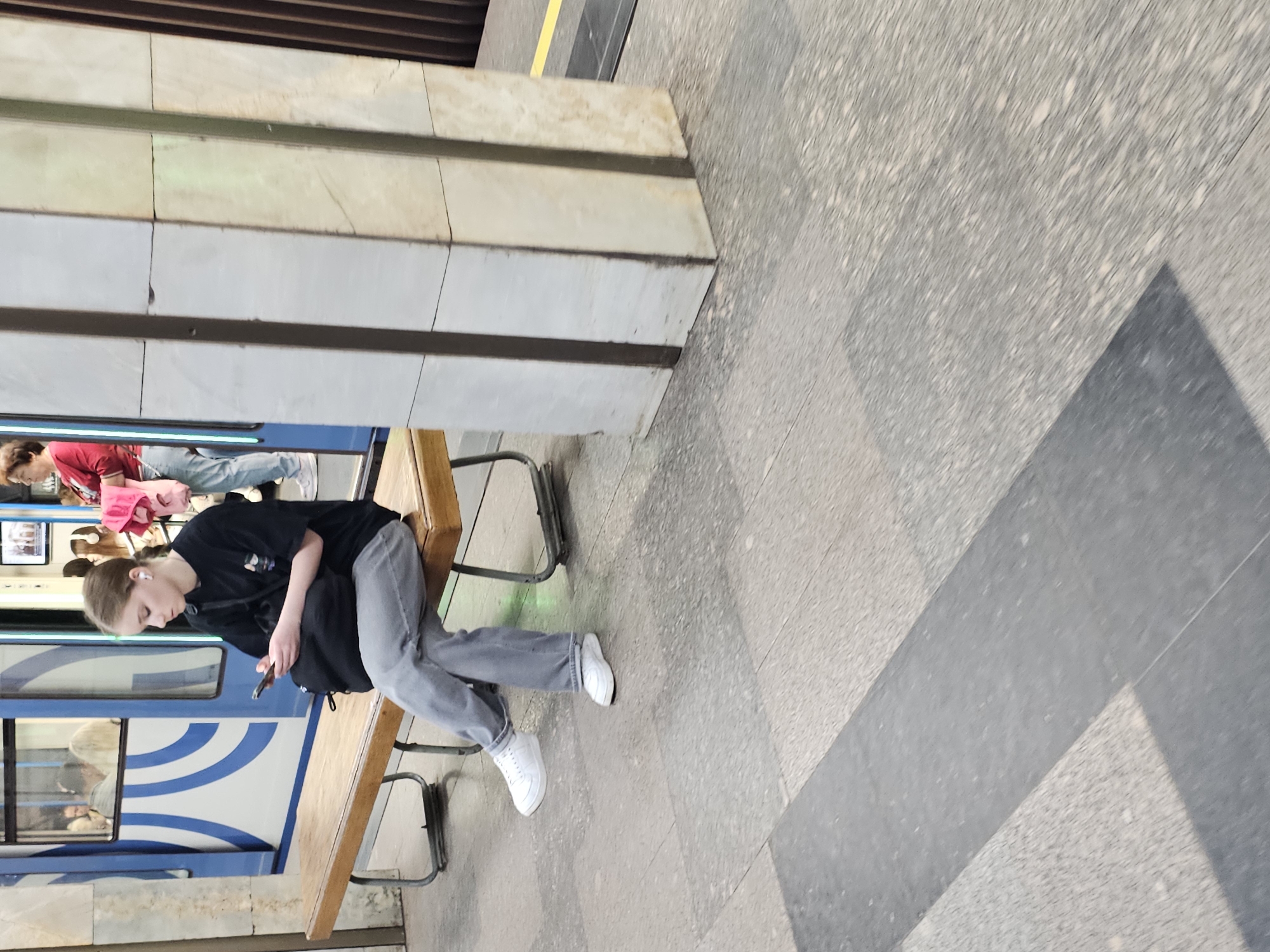 The image shows a person sitting on a wooden bench at a train station. The person is wearing a black t-shirt, grey pants, and white shoes. They are looking at a mobile phone in their hands. A train car with blue and white stripes is visible behind the person. Other people are visible in the background, partially obscured. The floor is grey with darker grey geometric shapes, and a wall with horizontal lines is in the upper left corner. A yellow line is on the top of the frame. The location is a train station in the city of Unknown, Unknown.