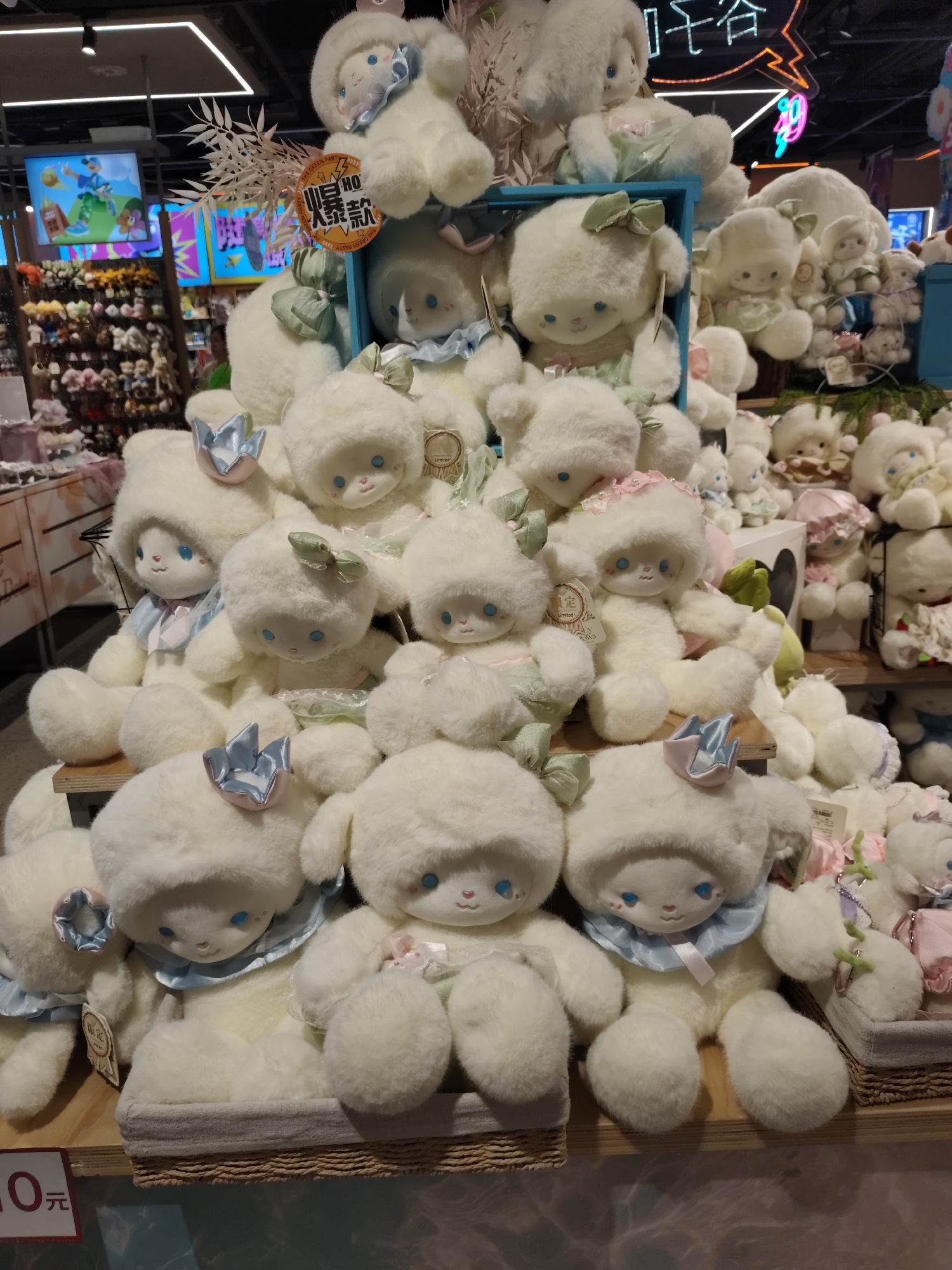 The image depicts a close-up of a shelf filled with numerous white, fluffy stuffed animals resembling sheep or lambs. They are arranged in clusters, with some smaller ones nestled behind larger ones. Many of the stuffed animals have blue eyes and wear decorative bows or collars in pastel colors like light blue and green. Some have delicate crowns on their heads. The stuffed animals are placed on wooden shelves, and in the foreground, there's a red sign with white Chinese characters and the number "10". In the background, there are blurred displays of other merchandise and some neon signs, including one with prominent Chinese characters. The overall environment suggests a retail setting, likely a toy store or a section of a larger department store. The scene is well-lit, highlighting the soft texture of the stuffed animals. Given the sign with Chinese characters, this scene is likely located in China, and specifically in Daqing, China, based on the provided context.