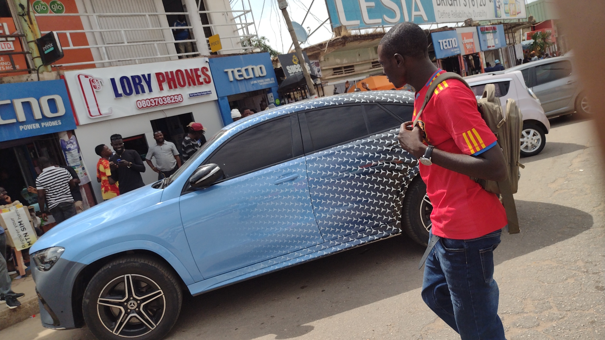 The image depicts a street scene in Jos, Nigeria, characterized by bustling commercial activity. A light blue Mercedes-Benz SUV with a distinctive star pattern paint scheme is parked on the side of the road. A man wearing a red shirt with yellow stripes on the sleeves, blue jeans, and a backpack walks past the vehicle. Various storefronts line the street, including "LORY PHONES" and "TECNO," both prominently displaying signage for mobile phone brands. Additional shops and pedestrians are visible in the background, indicating a busy urban environment. The sky is partly cloudy, suggesting it is daytime.
