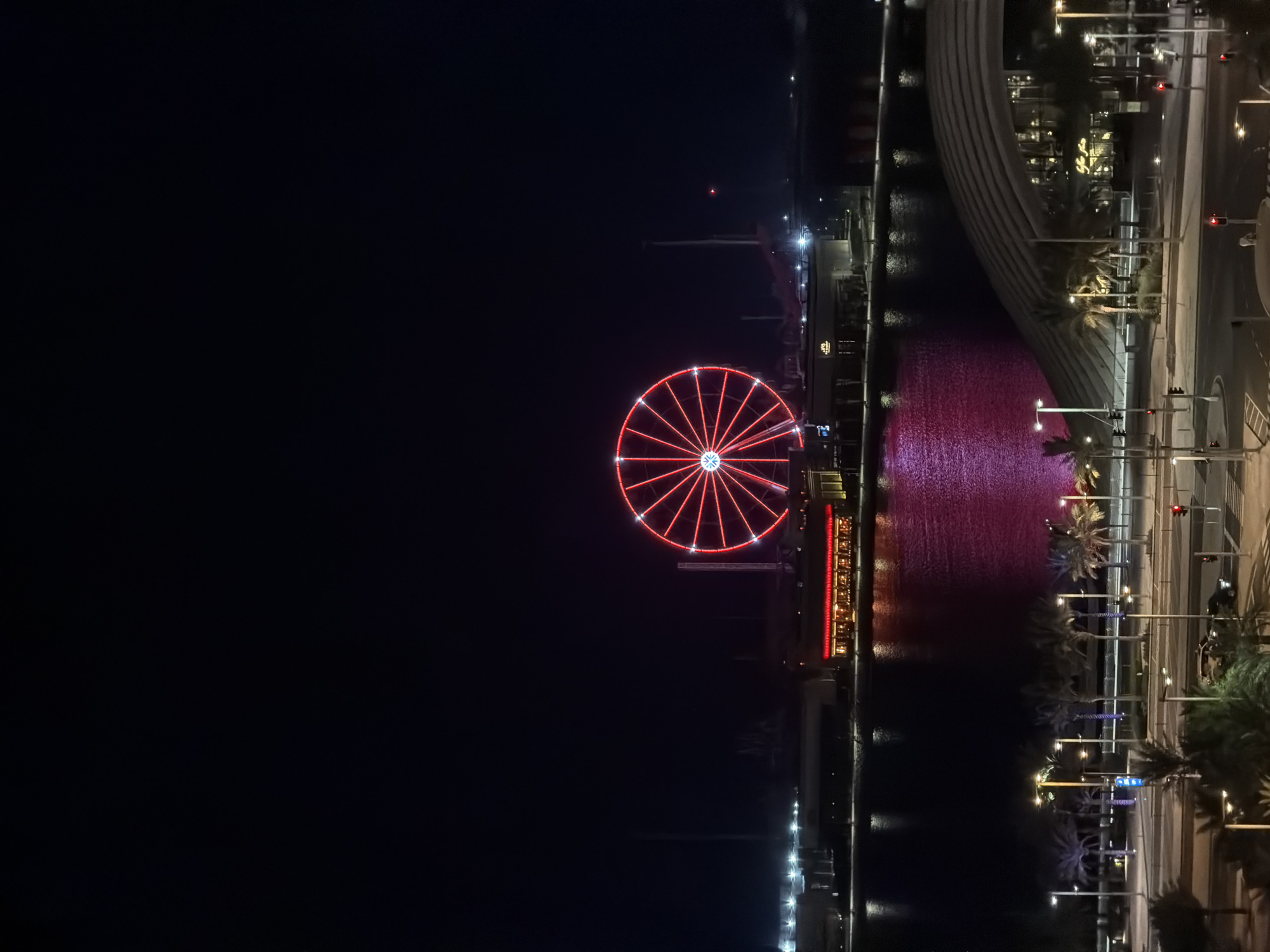 This image depicts a nighttime scene of a waterfront area in an unknown city. A large Ferris wheel, illuminated with red lights, is the central focal point. The Ferris wheel is situated across a body of water from the viewer. Along the waterfront, there are buildings with illuminated windows, suggesting commercial or residential structures. The water reflects the lights from the Ferris wheel and buildings, creating a pinkish hue. In the foreground, there is a street with traffic lights and palm trees. A modern, curved building with illuminated windows is visible on the right side of the foreground. The sky is dark and clear.