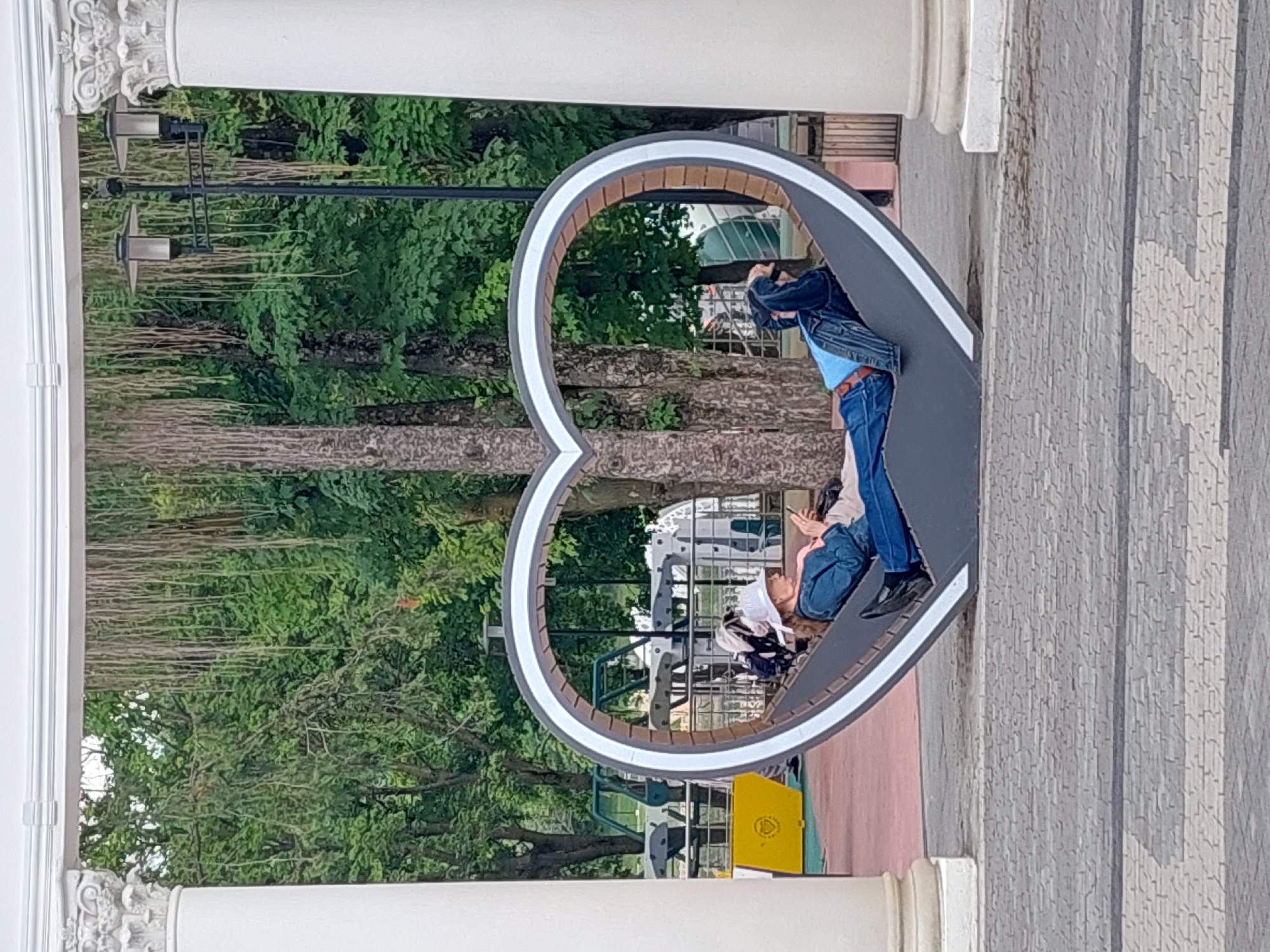An unidentified person, wearing a white hat and blue denim jacket over a pink shirt, reclines on a grey, heart-shaped bench. The person is holding a mobile phone and appears to be looking at it. The bench is situated outdoors, framed by two white columns and an archway, with a park-like setting in the background featuring trees and foliage. In the distance, there are some playground-like structures and a lamppost. The ground in front of the bench is paved with grey interlocking stones. The scene appears to be a public space, possibly a park or recreational area, in Alikonovka, Russia.