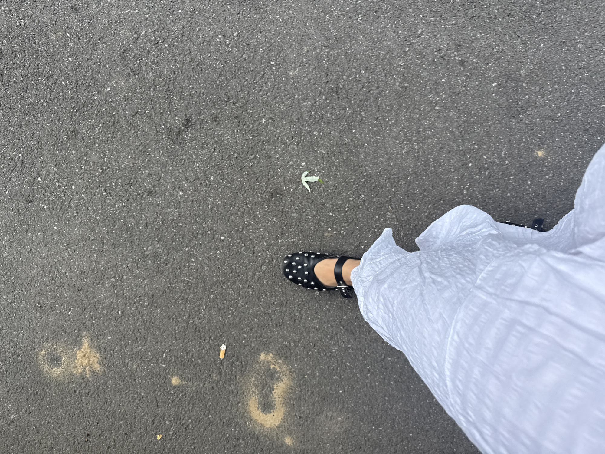 A high-angle, downward-facing perspective captures the lower portion of an individual, likely standing or walking on asphalt pavement. The visible leg, partially obscured by a white, crinkled fabric garment, terminates in a black, studded Mary Jane-style shoe with a buckled strap. The shoe features numerous small, metallic studs across its surface. The pavement, consistent with urban infrastructure in Milan, Italy, is dark grey asphalt with visible aggregate. Scattered on the asphalt are two cigarette butts, one positioned towards the upper left and another in the lower central area of the left side. A small, light green leaf, shaped somewhat like an arrow pointing upwards, rests on the asphalt in the middle right portion of the frame. Additionally, faint, irregular yellowish-brown stains or markings are visible on the asphalt, particularly in the upper left quadrant and below the person's leg.