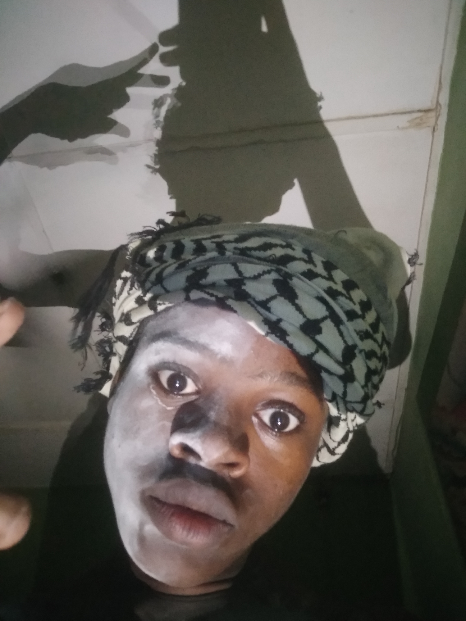 A close-up, low-angle shot of a person wearing a black and white keffiyeh. The person's face has a white substance on it, possibly cream or paint. The background consists of a white ceiling with the shadow of a hand and arm. Part of a green wall is visible on the right side of the frame. The location is presumed to be Shangisha, Nigeria.