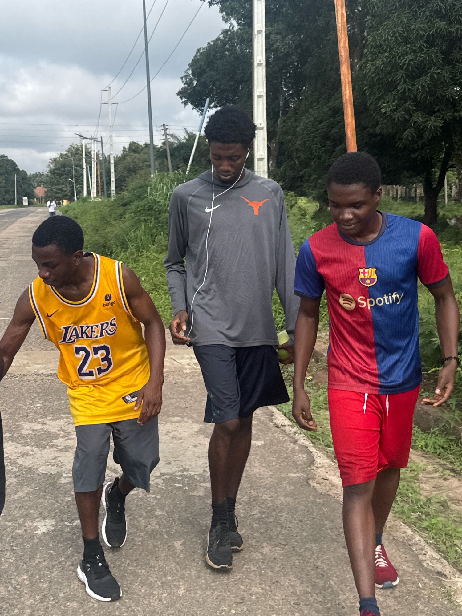 Three young Black men are walking on a paved road in Ihiagwa, Nigeria. The man on the left wears a yellow Los Angeles Lakers basketball jersey with the number 23. The man in the middle wears a gray long-sleeved shirt with an orange University of Texas logo and dark shorts. The man on the right wears a blue and red two-toned t-shirt and red shorts. All three men are wearing athletic shoes and socks. They are walking away from the camera, heading towards the background where trees and utility poles are visible. The sky is overcast, suggesting a cloudy day. The road appears to be a rural or semi-urban street, with vegetation lining one side and power lines overhead.