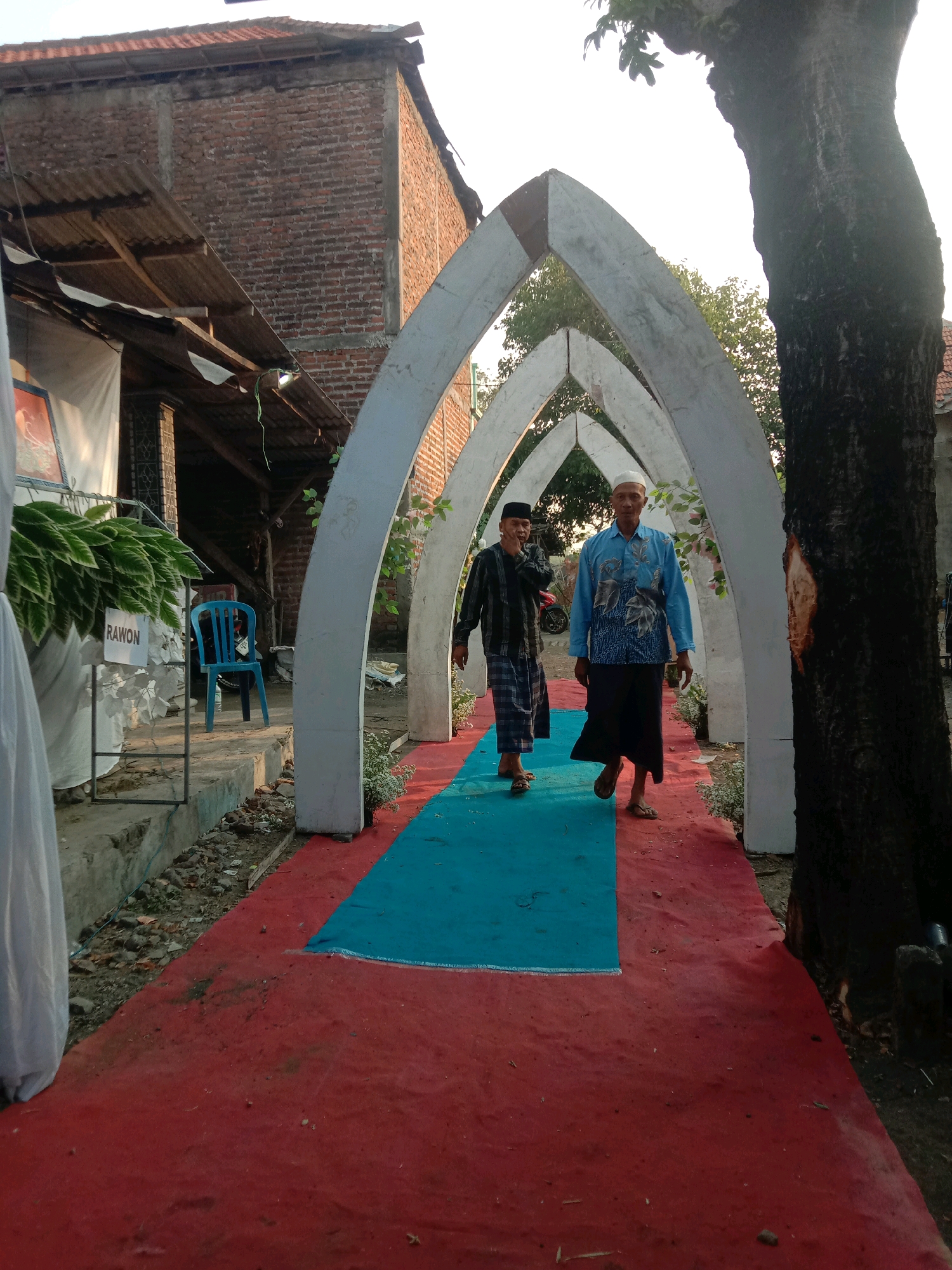 In a scene set in Karang Anyar, Indonesia, two men are walking under a white arched structure. The man in front is wearing a blue, long-sleeved shirt with a floral pattern and a dark sarong. He has a white skullcap on his head and appears to be looking forward. The man behind him is wearing a black and white checkered shirt and a plaid sarong, also with a white skullcap. He is looking to his left and has his hand raised near his mouth. A red carpet runs along the ground, bordered by a blue carpet in the middle of the walkway. The walkway is lined with small plants. To the left of the walkway, a blue plastic chair is visible next to a sign that reads "RAWON". Behind the chair, a white sheet is draped. The background features a brick building with a corrugated metal roof and a large tree trunk on the right side of the image. The lighting suggests it is late afternoon.
